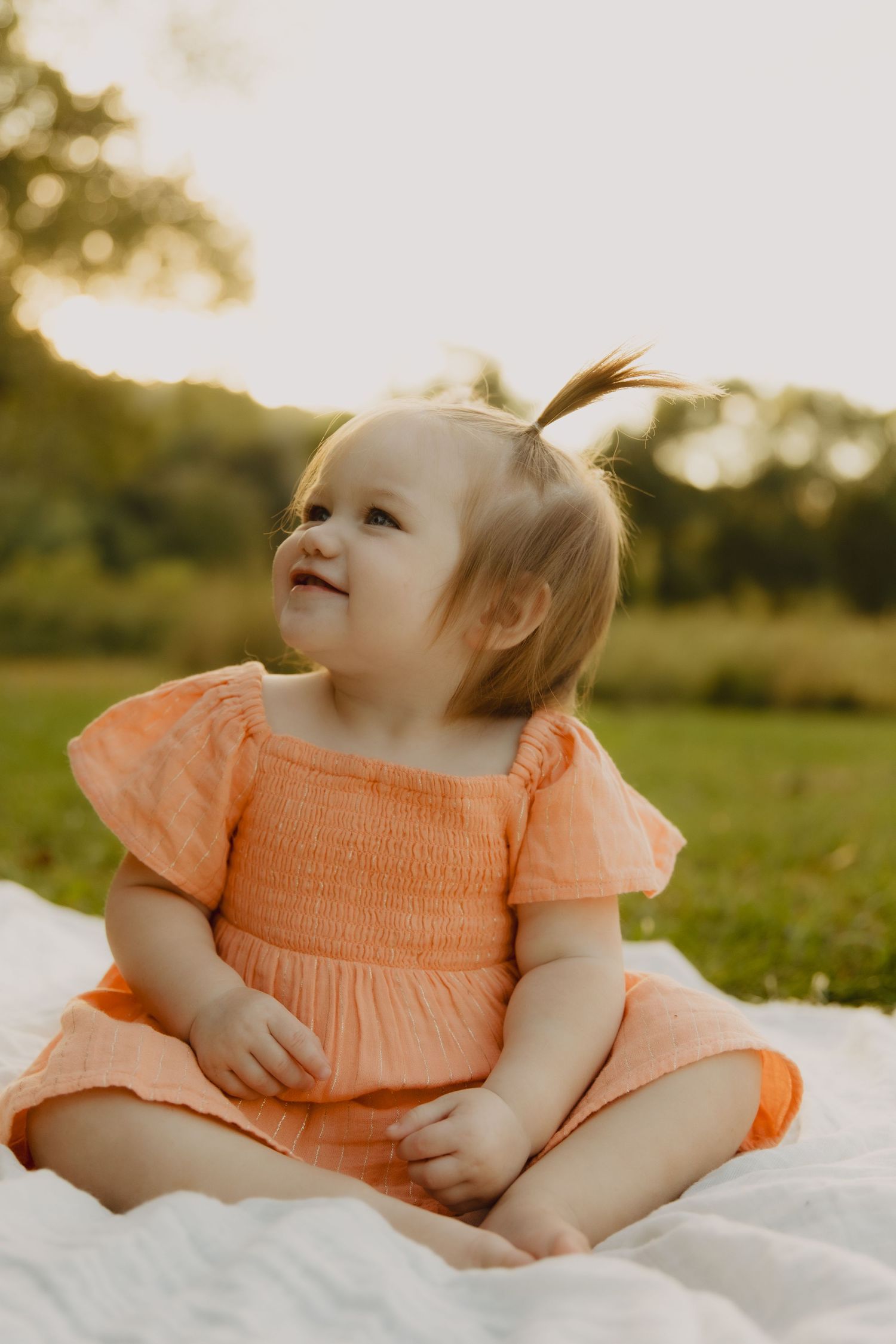 A young child in a peach-colored dress sits on a white blanket outdoors during golden hour.