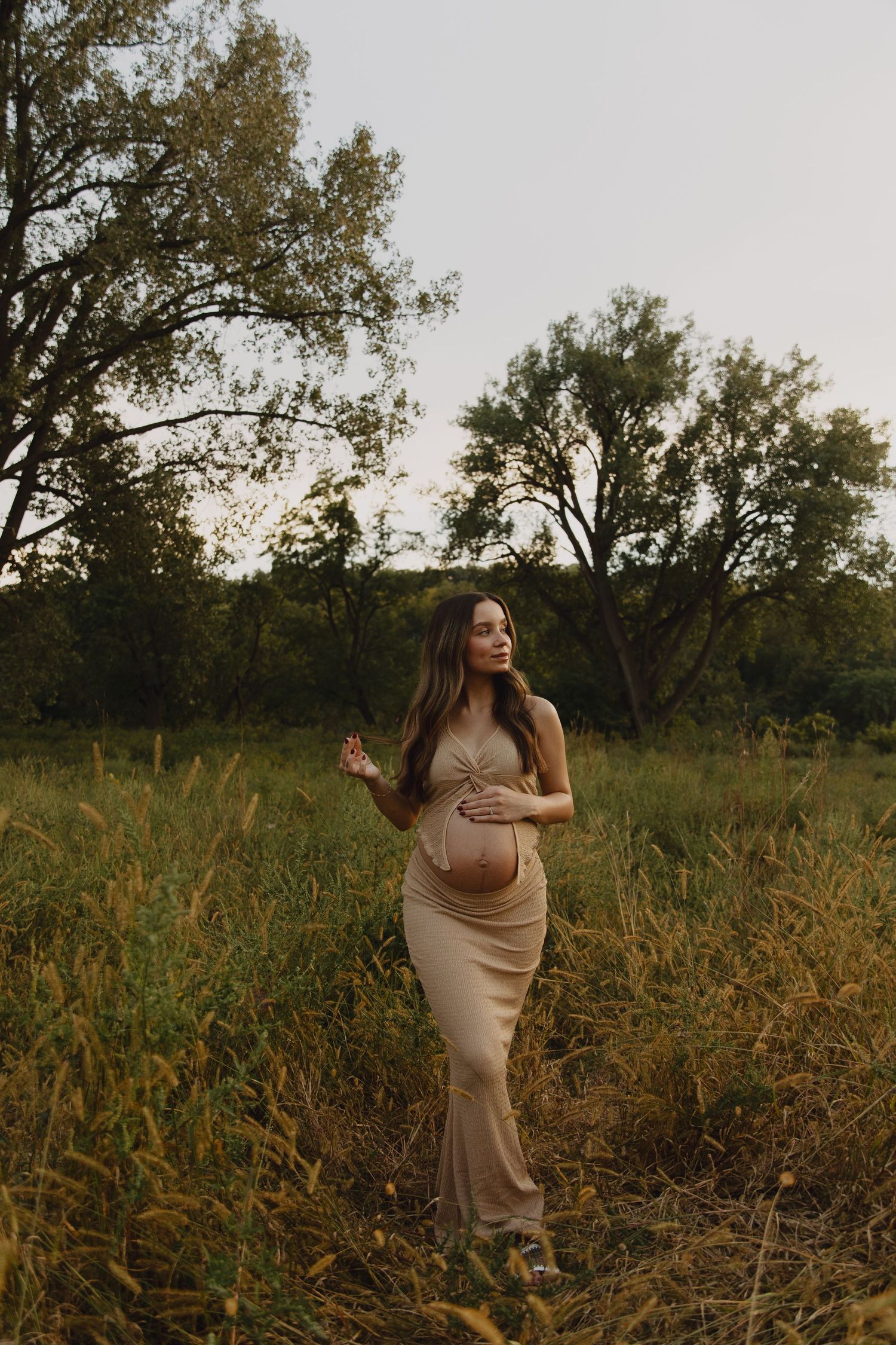 Pregnant woman in beige dress poses elegantly in natural outdoor setting.
