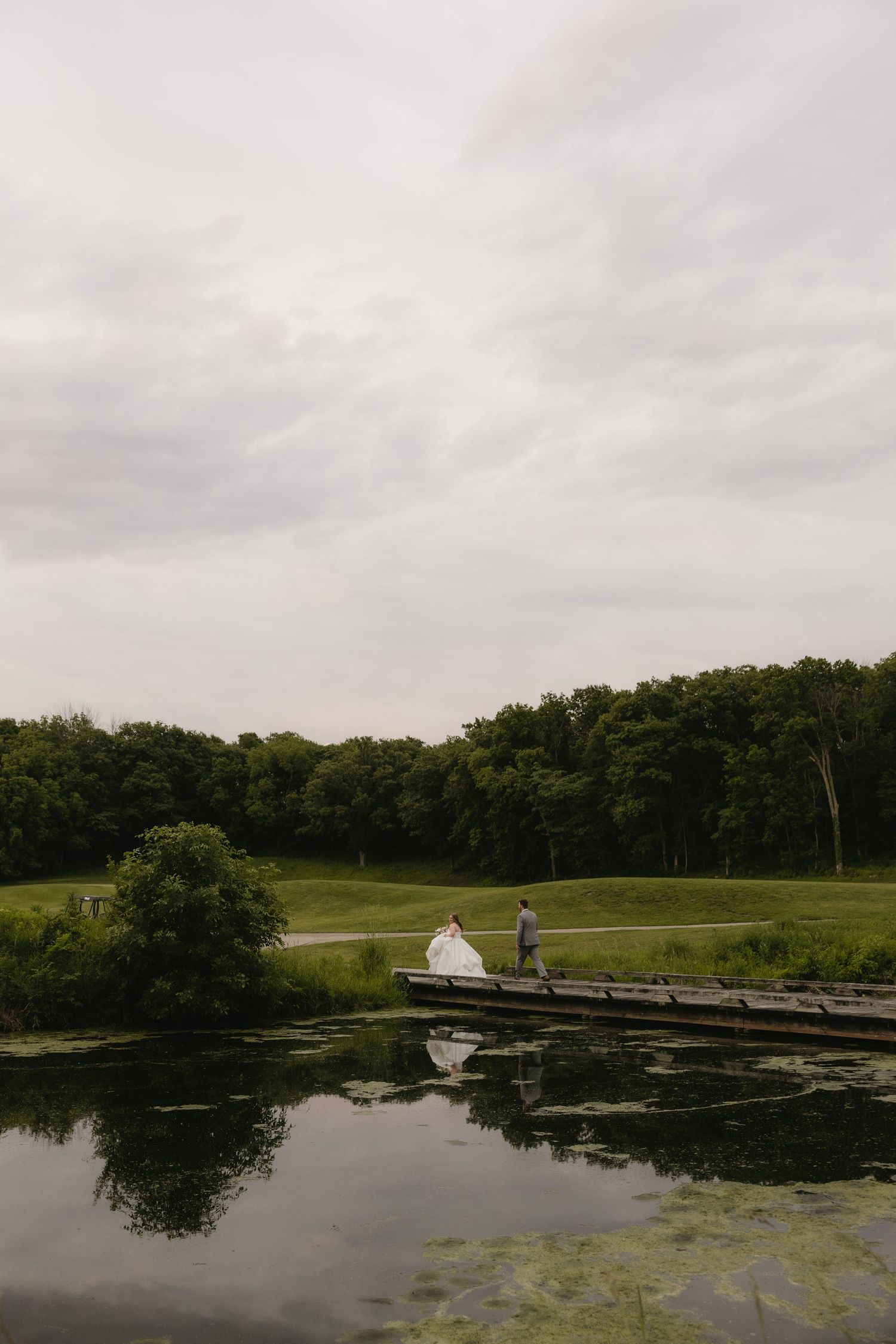 Bride and groom standing on a wooden dock by a reflective pond, surrounded by lush green trees under a cloudy sky.