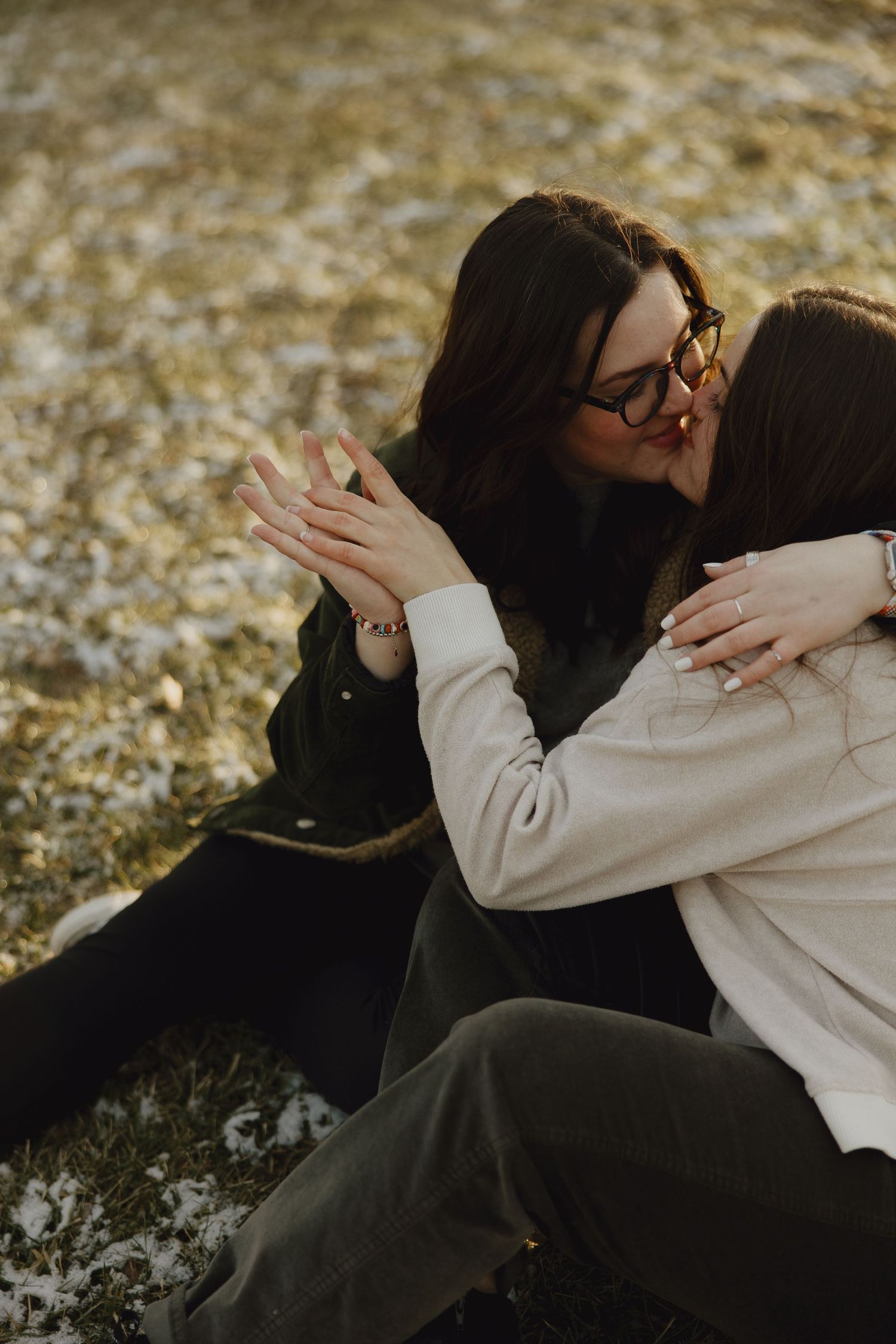Two people in warm clothing share an intimate embrace while sitting on grass during golden hour.