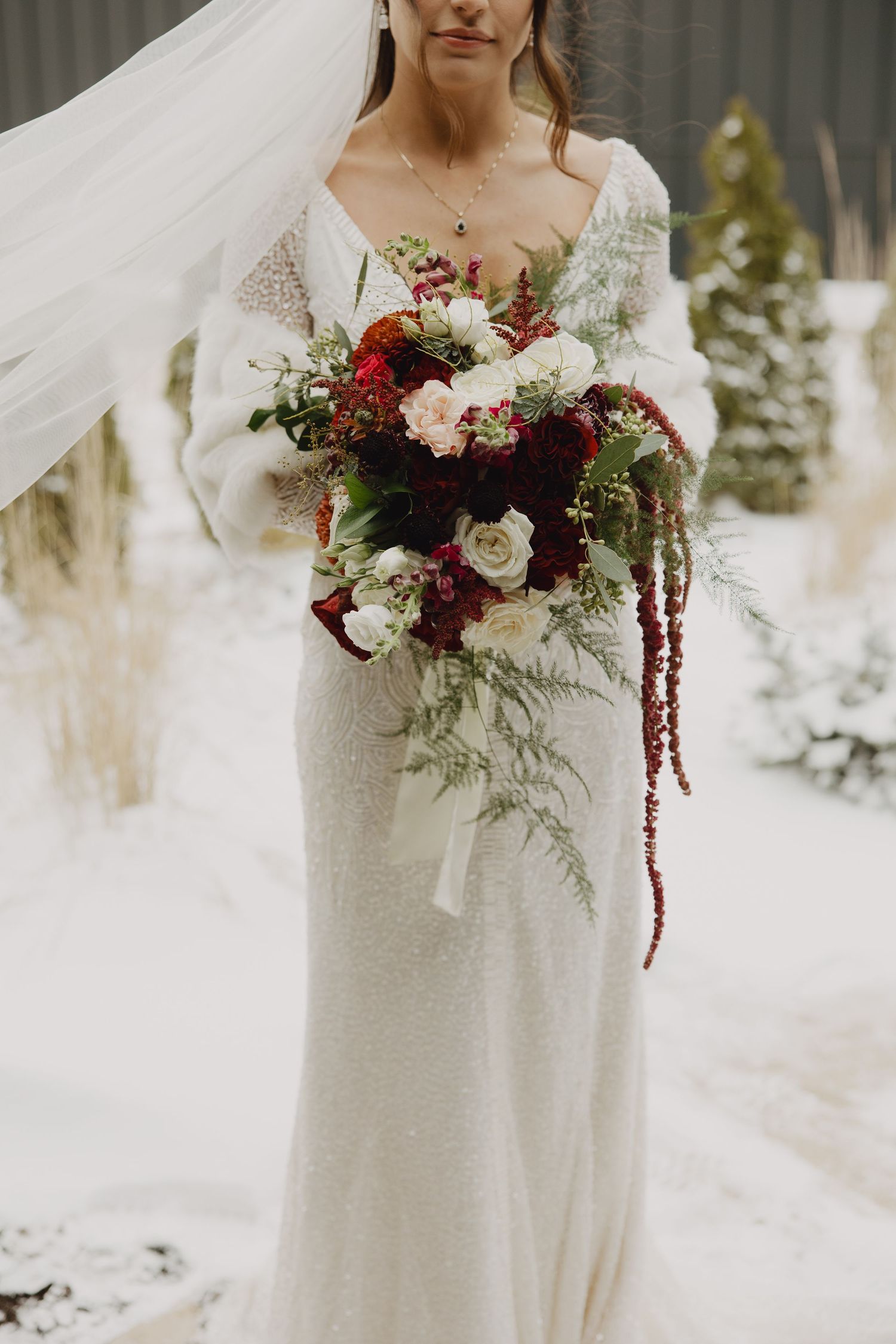 Bride in white sequin gown holding lush red and white winter bouquet with ferns in snowy outdoor setting.