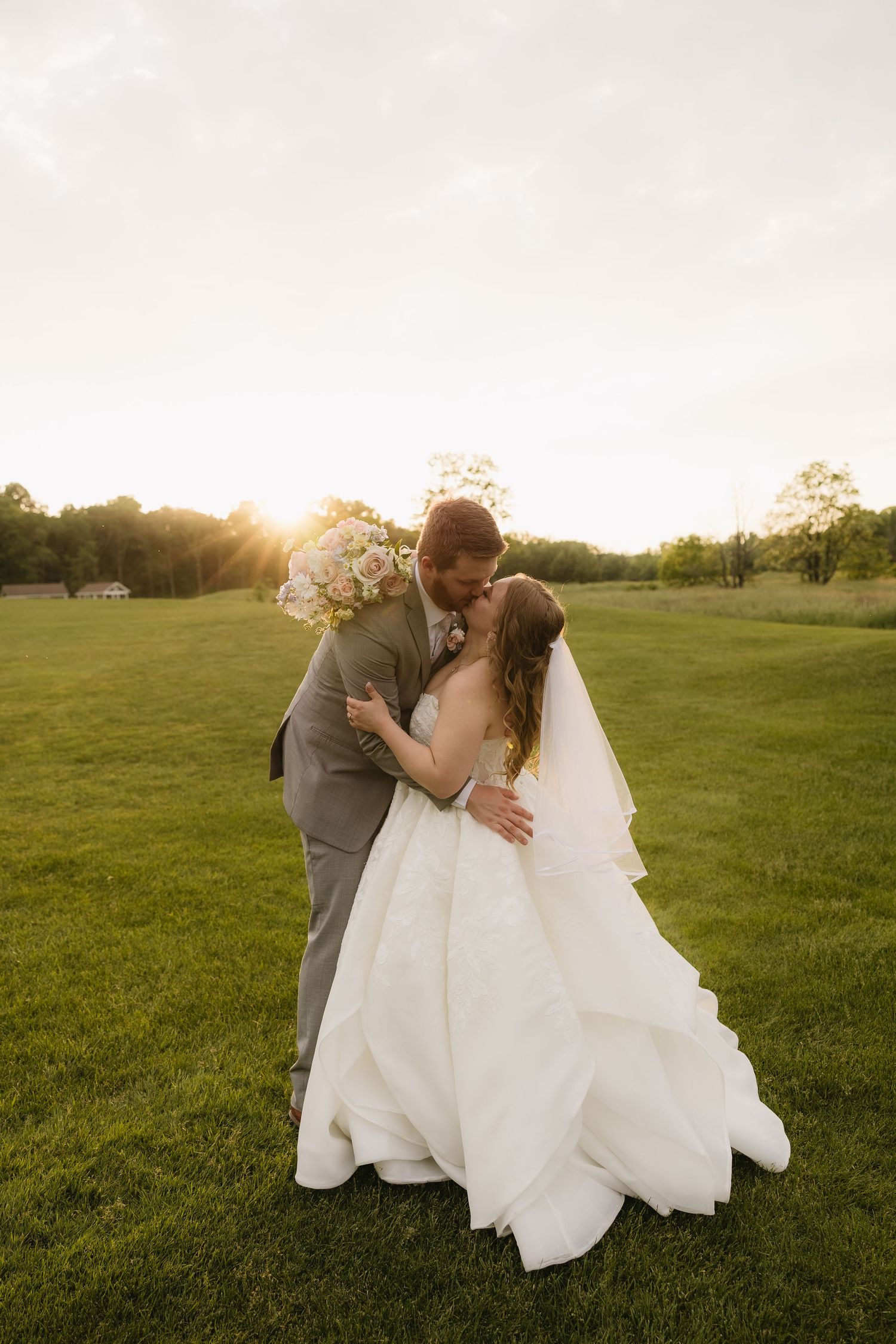 Bride and groom share a romantic kiss at golden hour on a lush green lawn, bride in white gown holding bouquet.