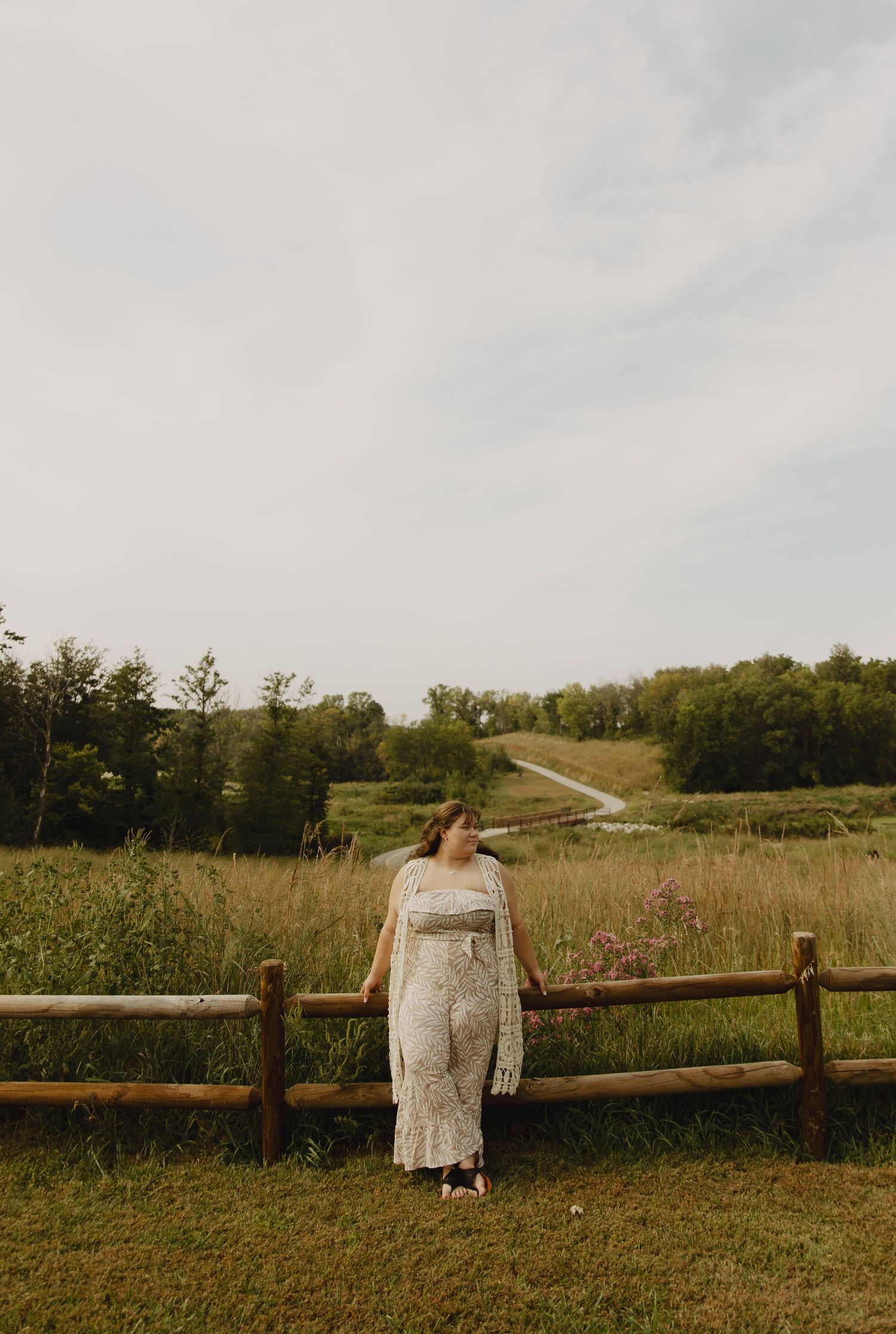 A person in a white dress stands by a rustic wooden fence overlooking rolling hills and forest on an overcast day.