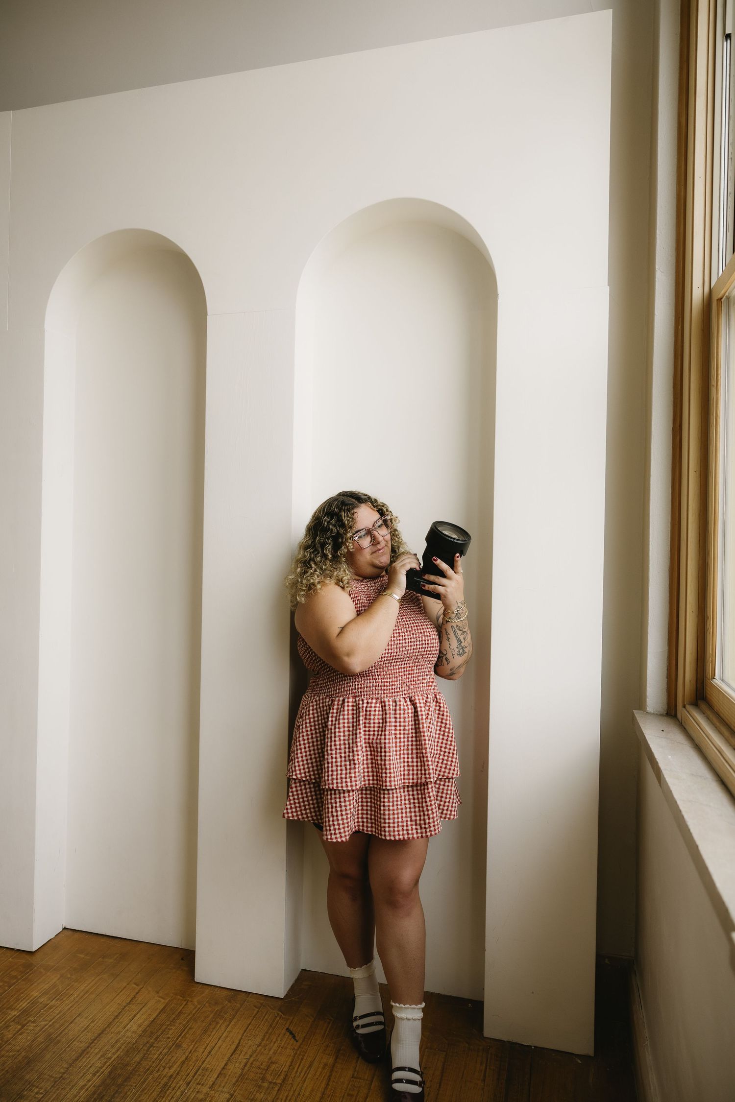 Woman in pink ruffled mini dress leaning against white arched wall alcoves, holding a camera, wearing white socks.