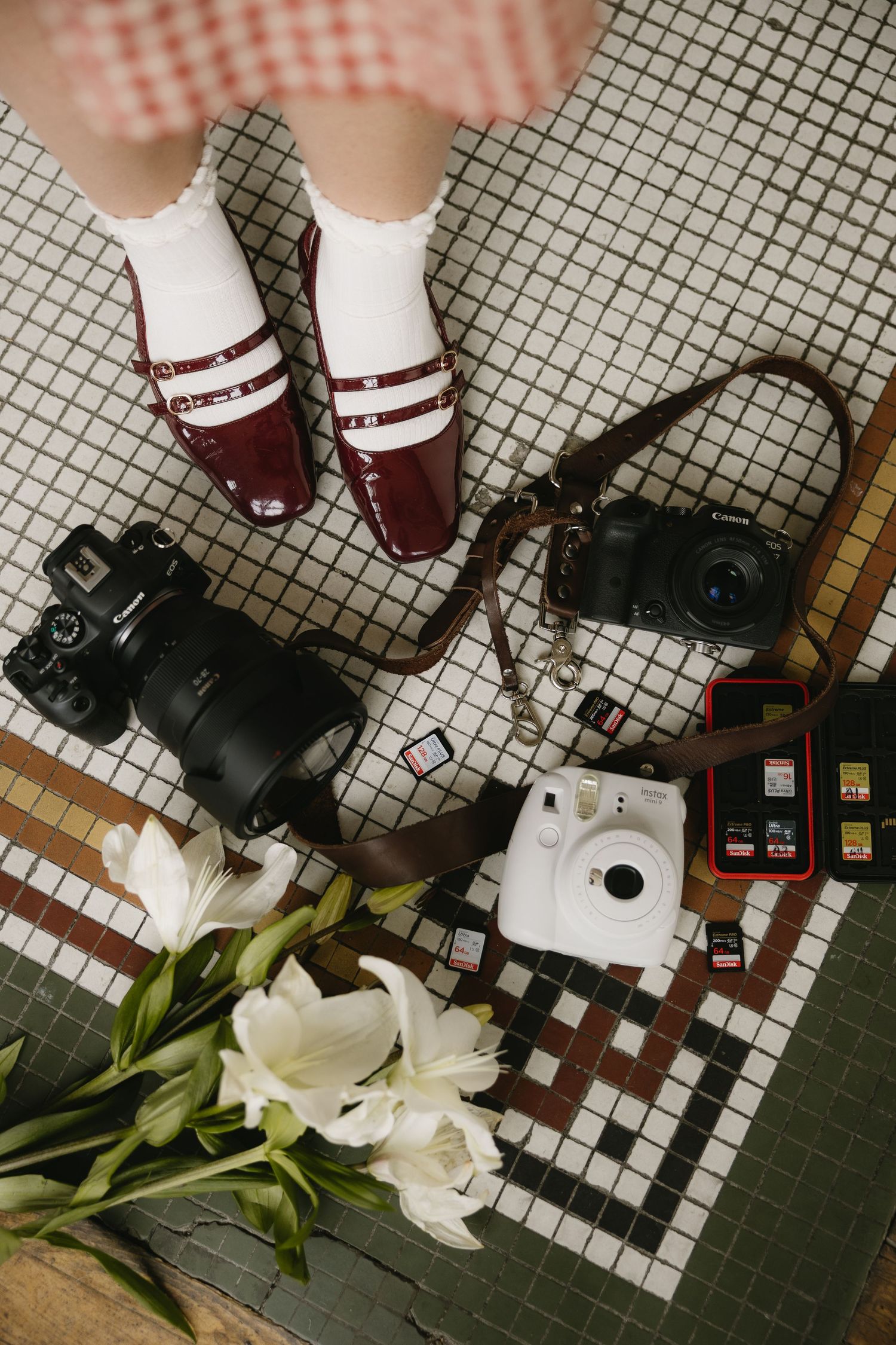 Flat lay of cameras, memory cards, white flowers, and burgundy loafers with white socks on a mosaic tile floor.