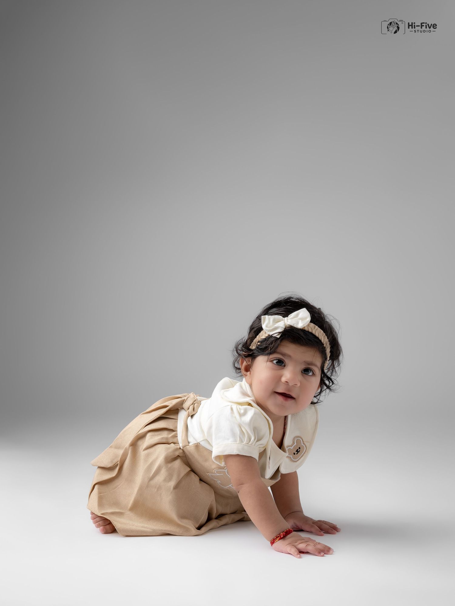 Cute Indian baby girl in cream and khaki dress with bow headband crawls in a professional studio setting.