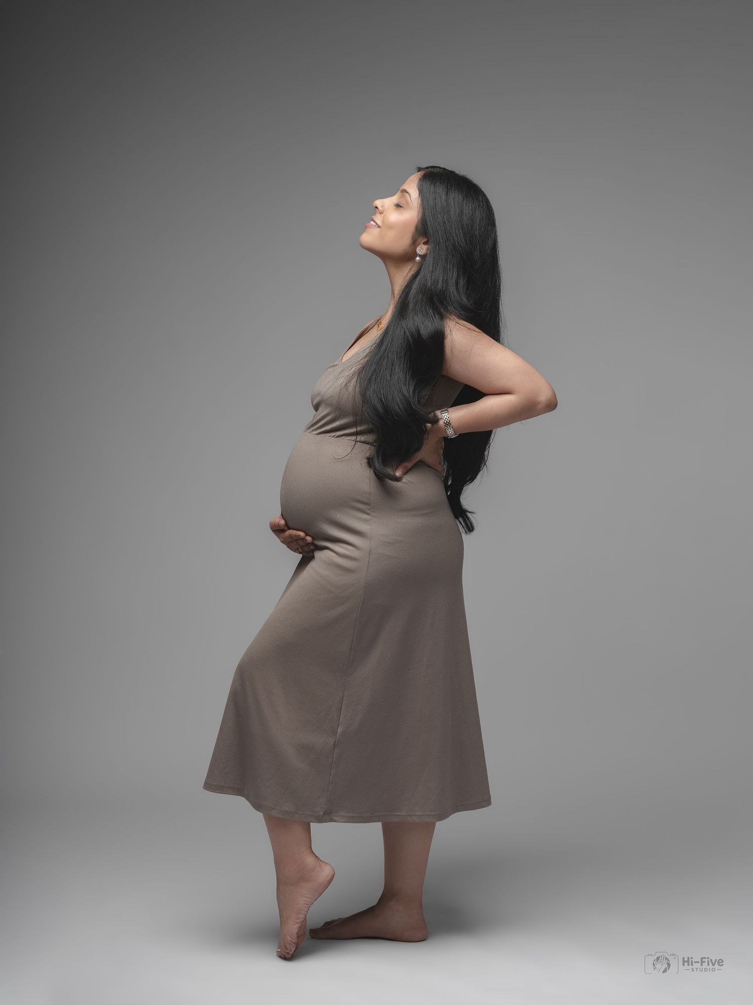 Side view of pregnant woman in taupe dress arching back gracefully during professional maternity shoot.