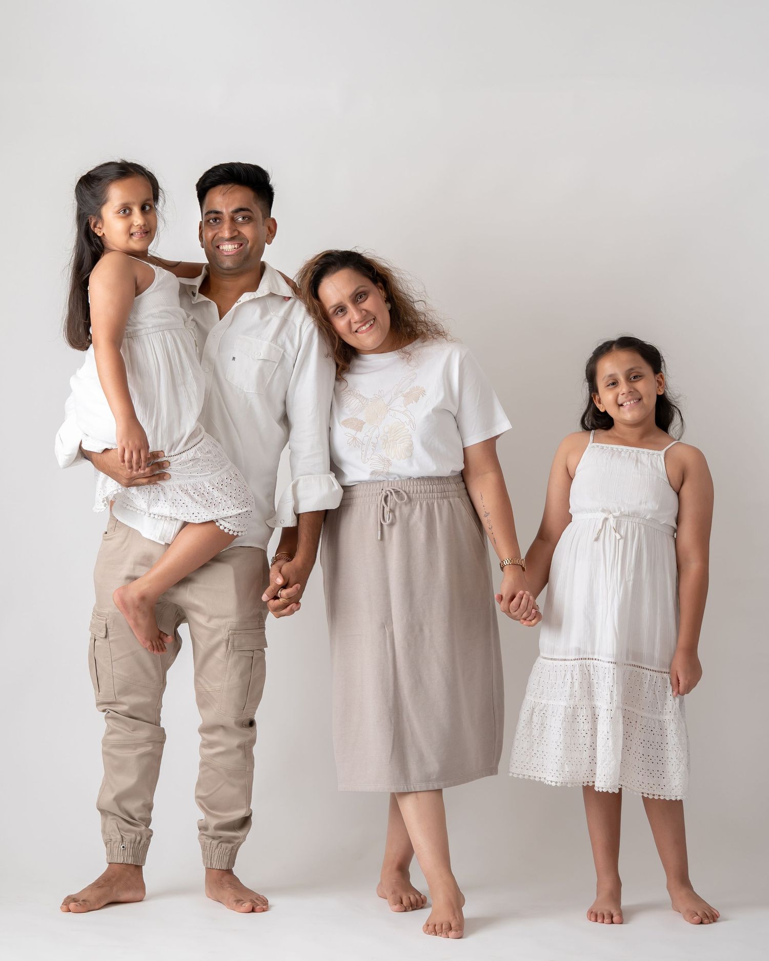 Family of four barefoot in white and beige outfits holding hands in a bright studio setting.