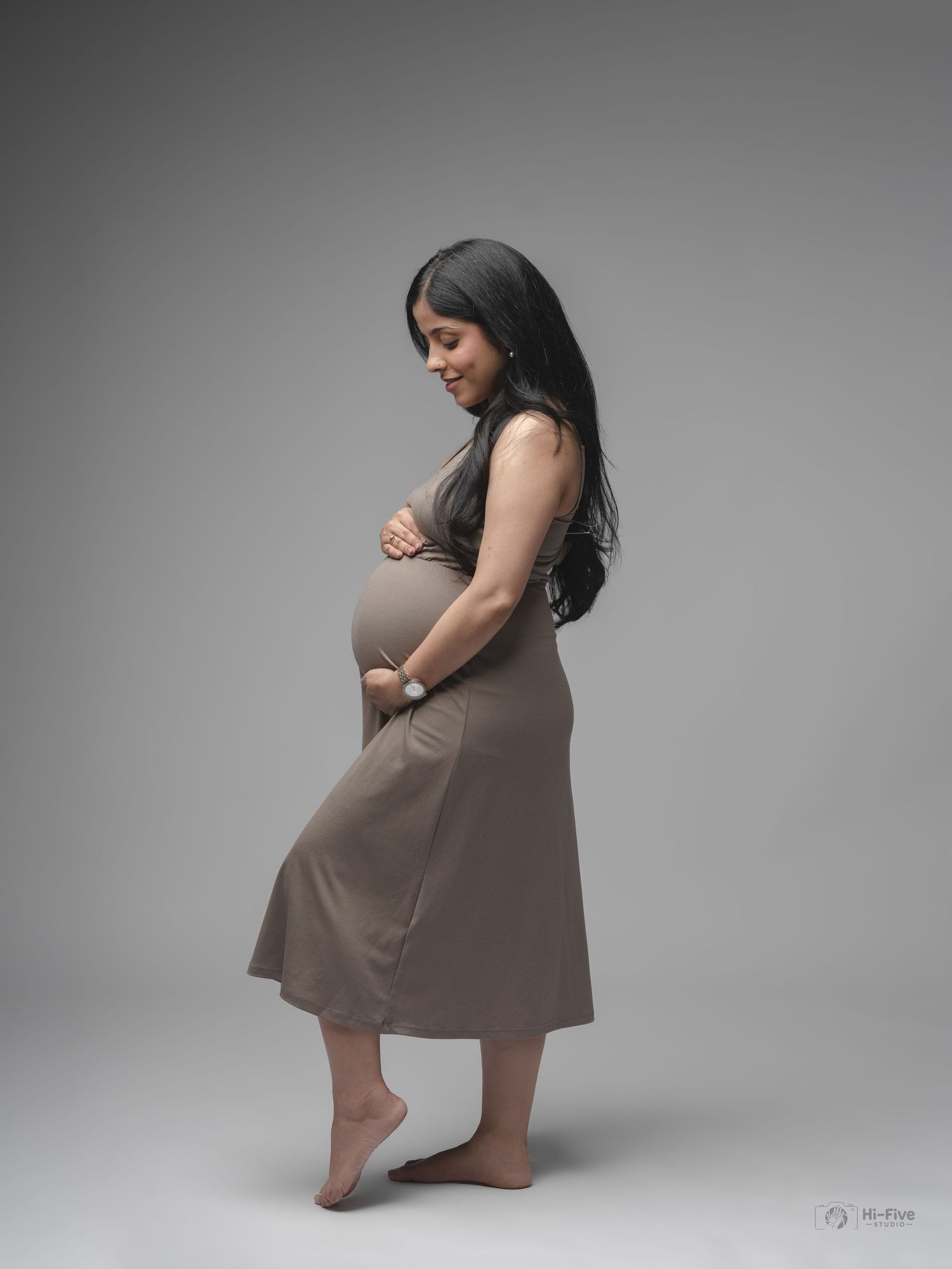 Pregnant woman in taupe dress cradling baby bump in elegant side profile studio shot.