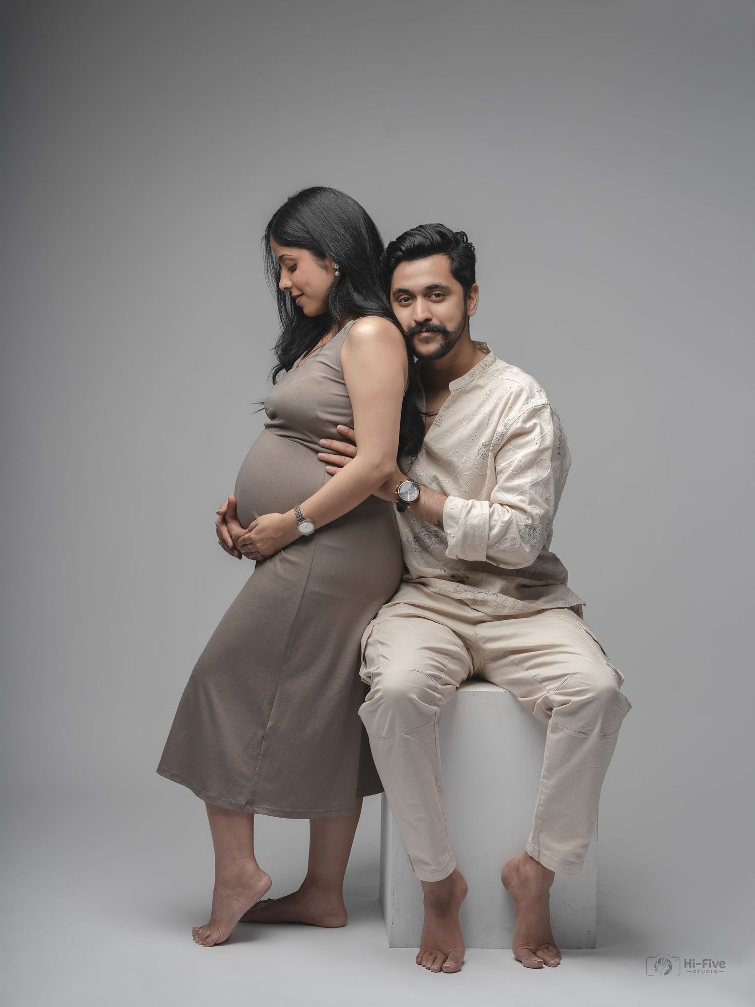 Maternity couple pose with pregnant woman standing beside seated partner against neutral studio backdrop.