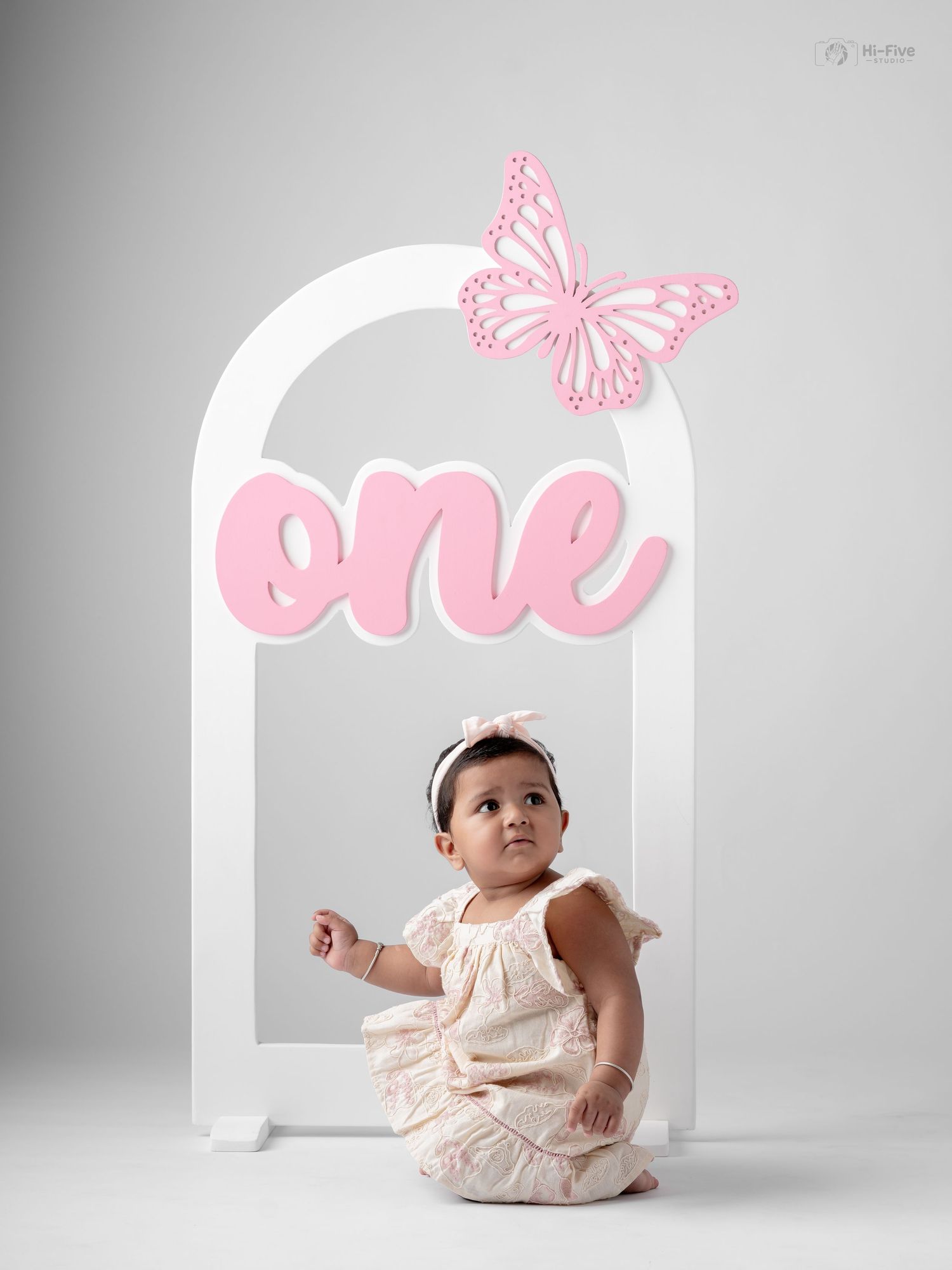 A baby girl in a cream dress sits before a white arch prop with pink “one” sign and butterfly for her first birthday.