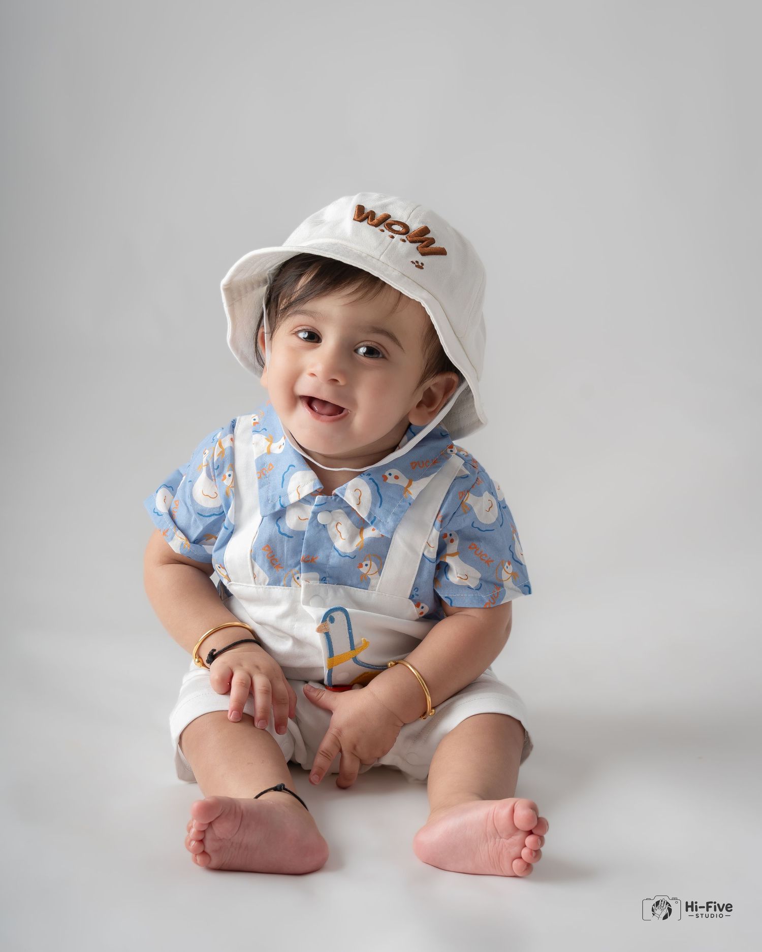 Smiling baby boy in blue duck print shirt and white bucket hat sitting on white background.