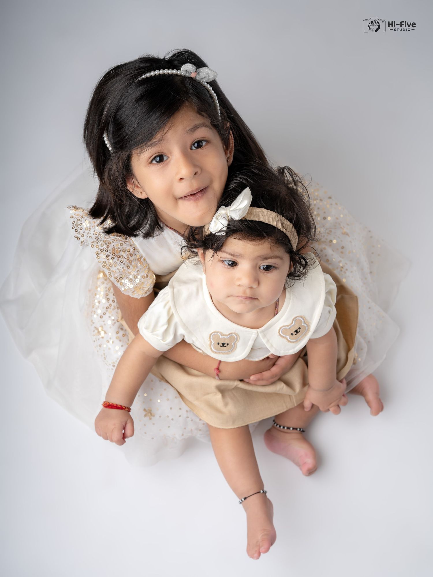 Overhead view of two Indian sisters in white and cream outfits embracing in a professional studio photo.