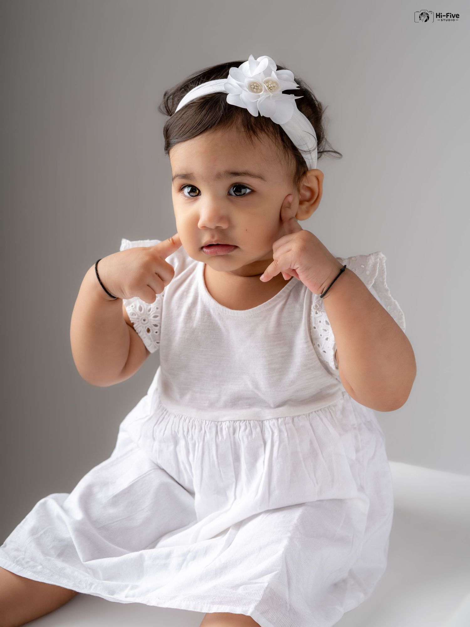 Adorable baby girl in white eyelet dress with bow headband posing with hands near face.