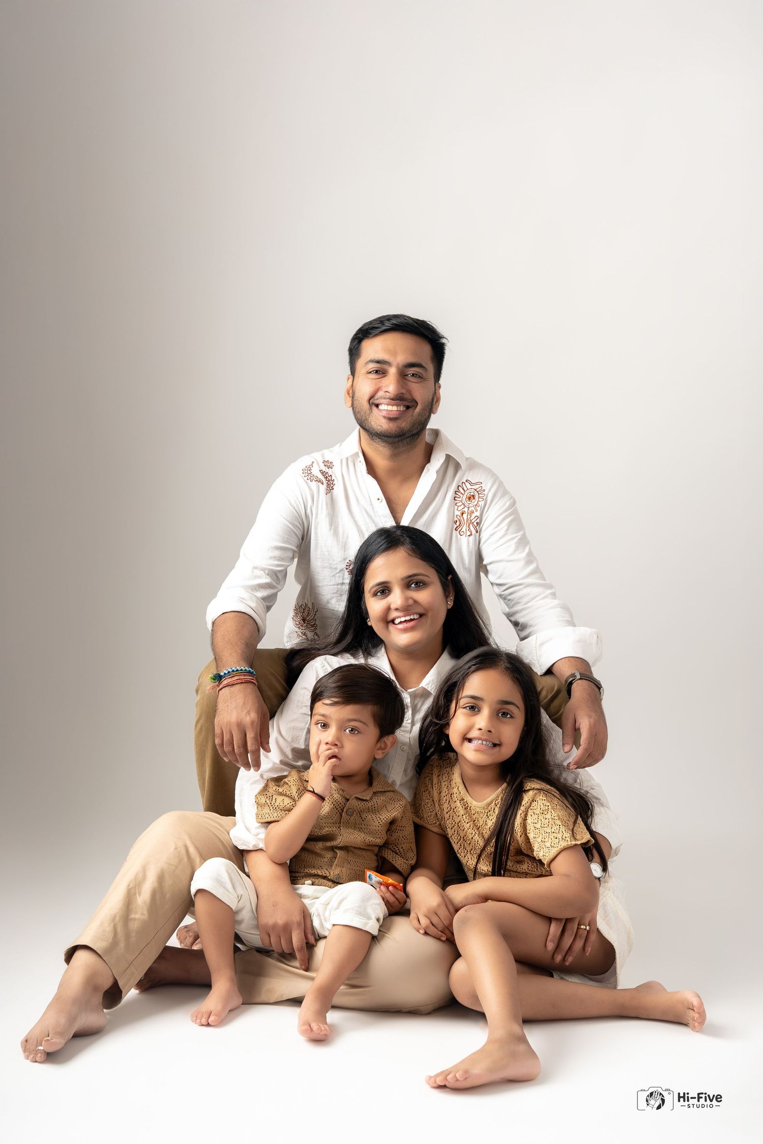 A smiling Indian family of four poses together in a studio, wearing white and beige coordinated outfits.