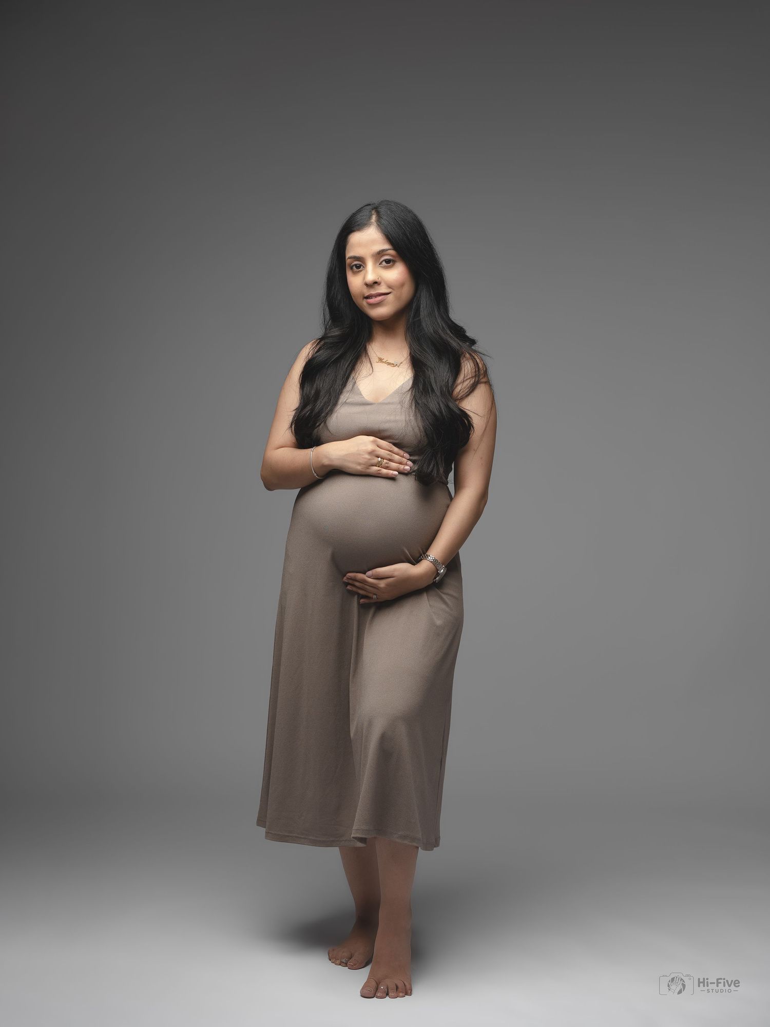 Expectant mother in fitted taupe dress smiling warmly while cradling baby bump in studio portrait.