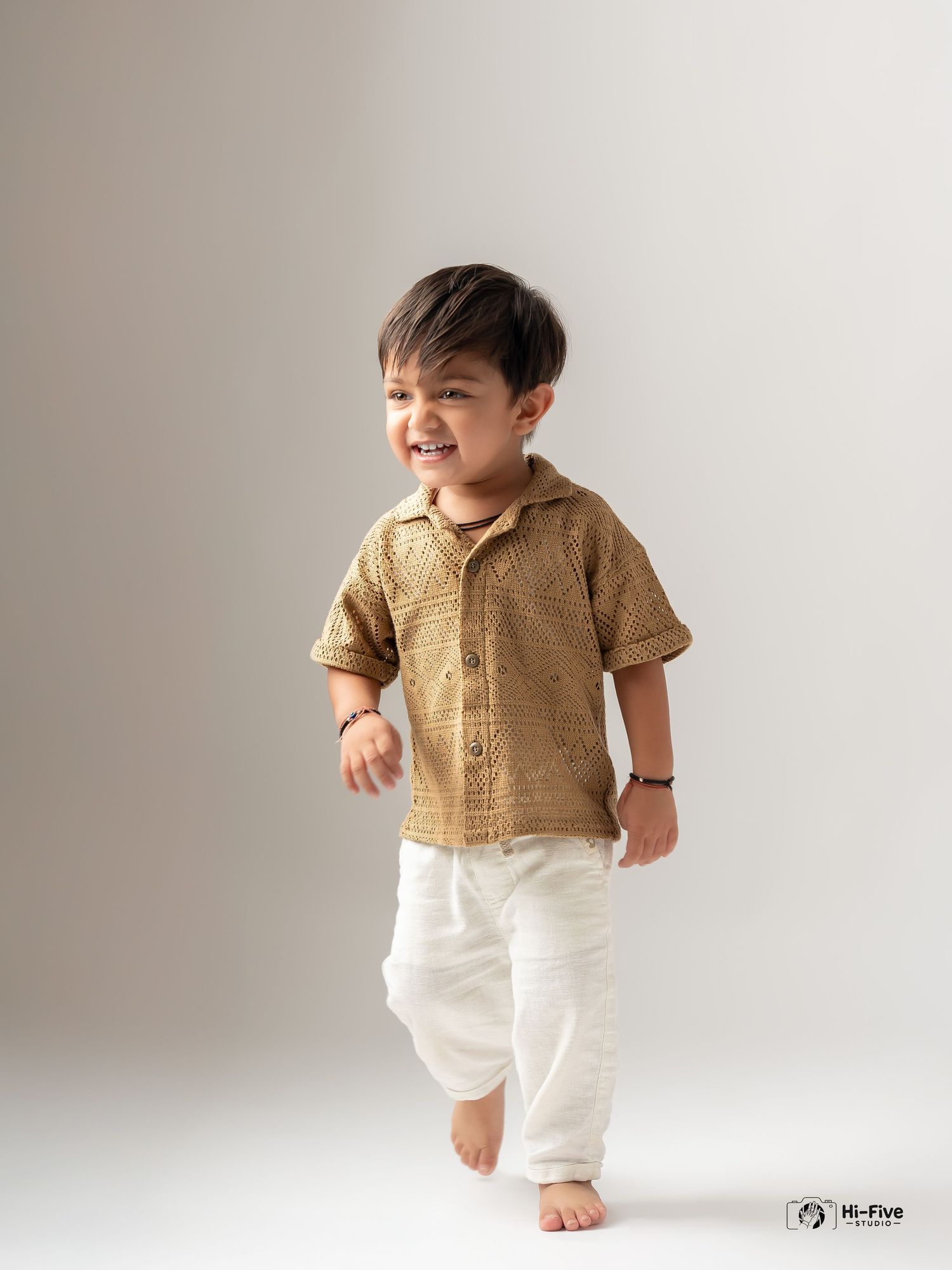 Happy toddler boy in gold printed shirt and white pants walking barefoot in studio.