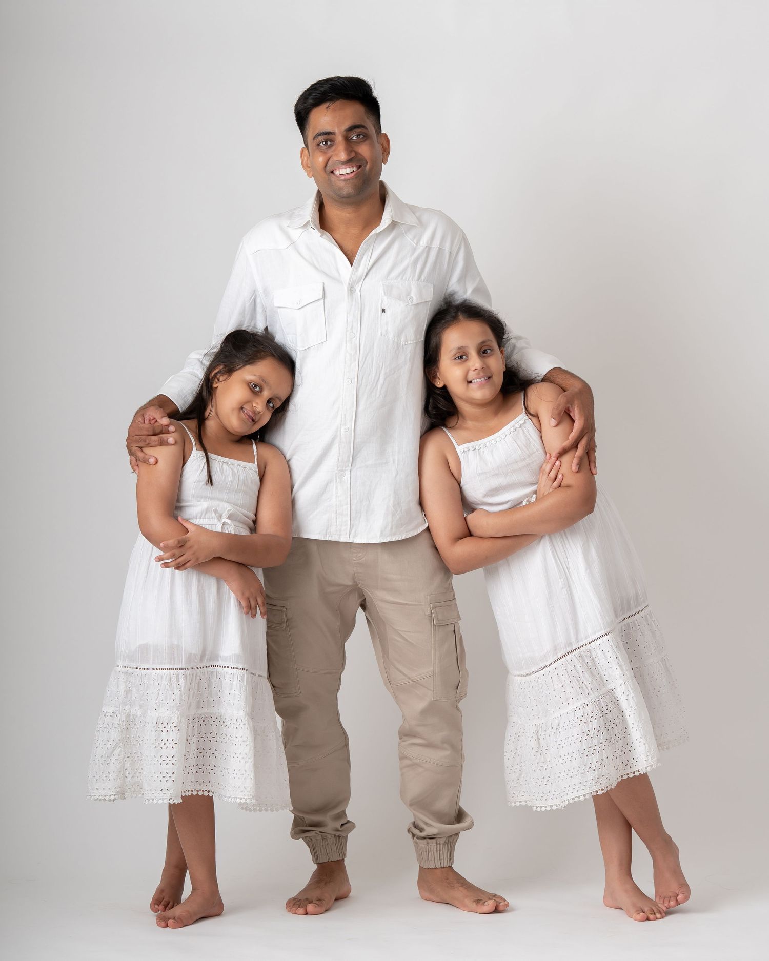 A smiling man in white shirt with two young girls in white dresses posing together for a family portrait.