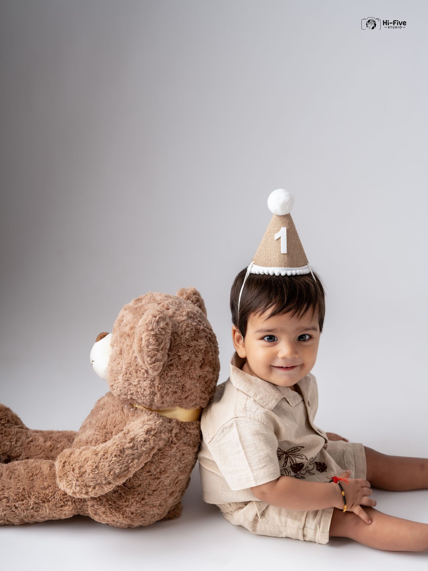 A smiling baby boy in a number one birthday hat poses with a large teddy bear in a studio.