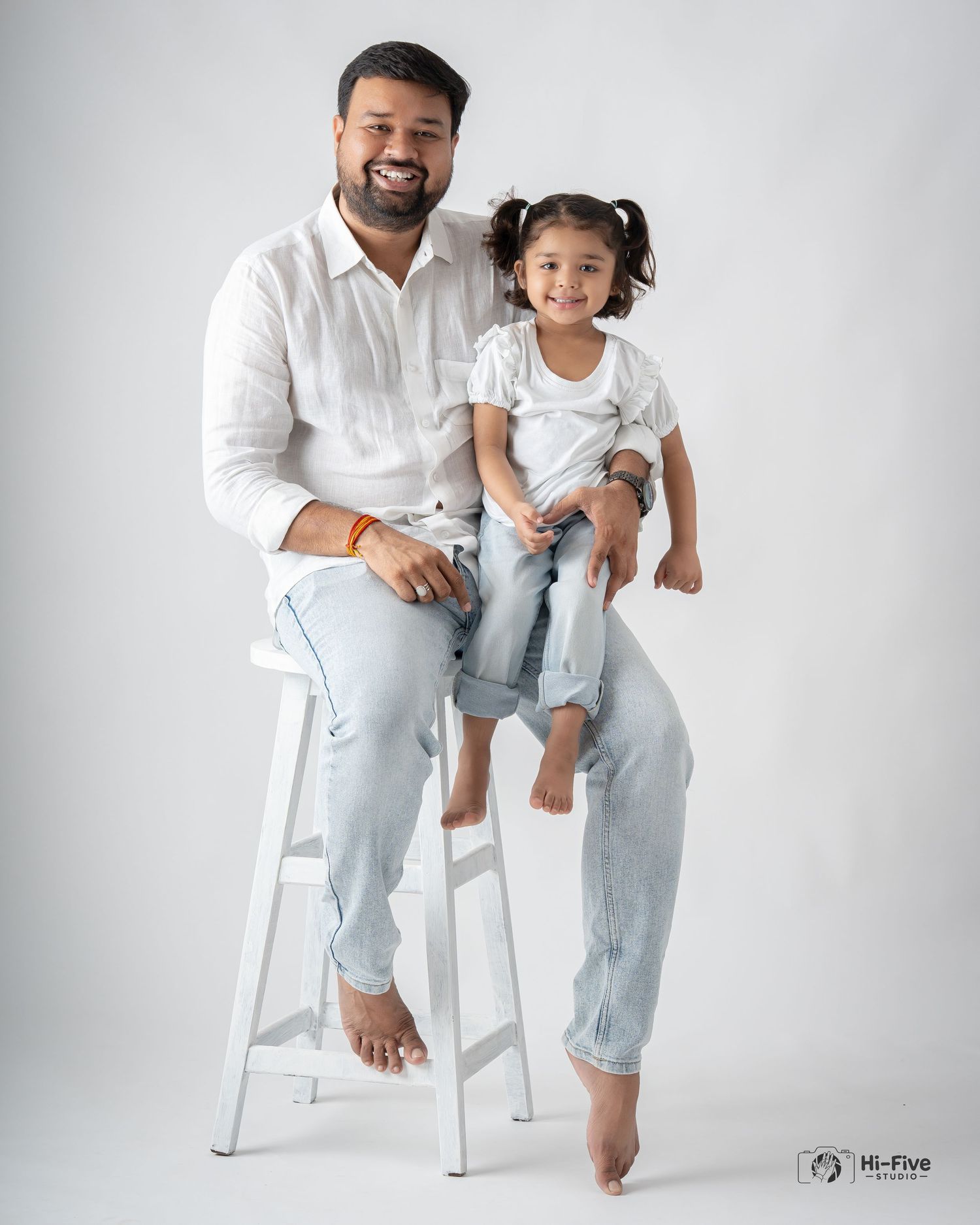 Smiling father and young daughter in white and light blue jeans posing on a stool in a studio.