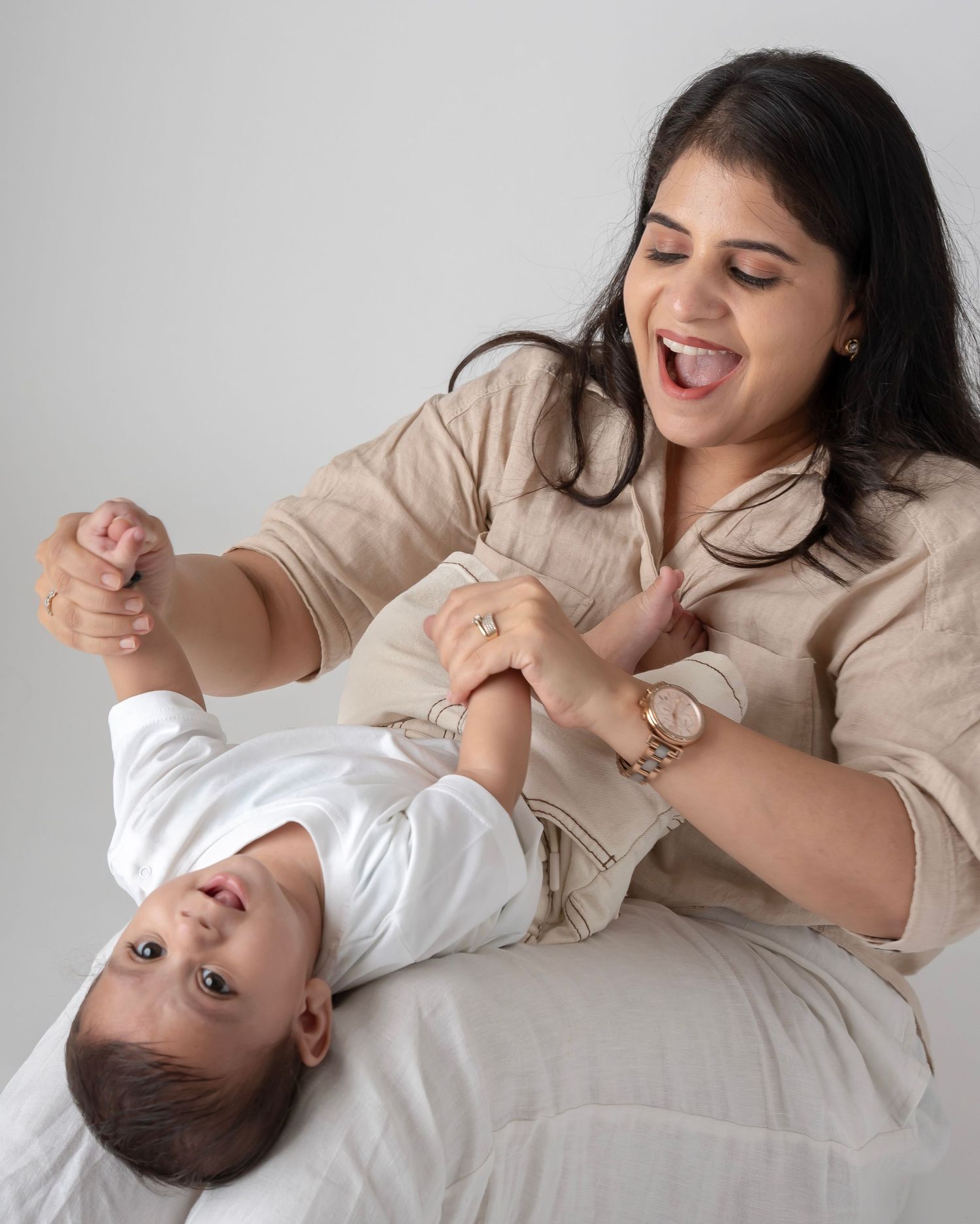 A laughing mother in beige shirt playfully holds her baby upside down during a fun studio photo session.