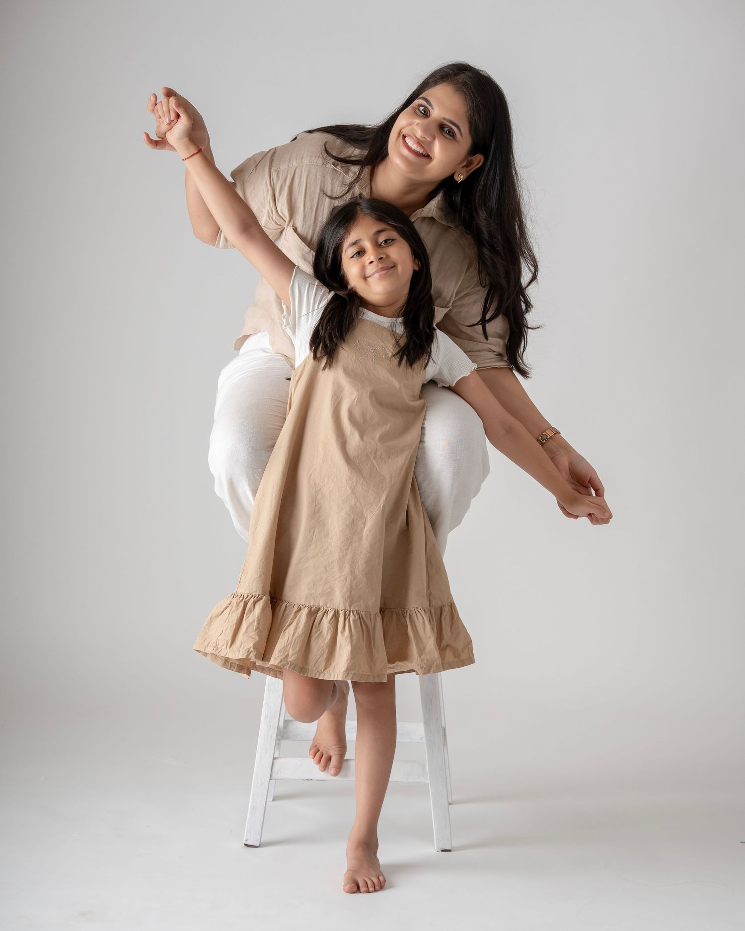 A mother and daughter pose playfully in matching beige outfits against a white studio background.