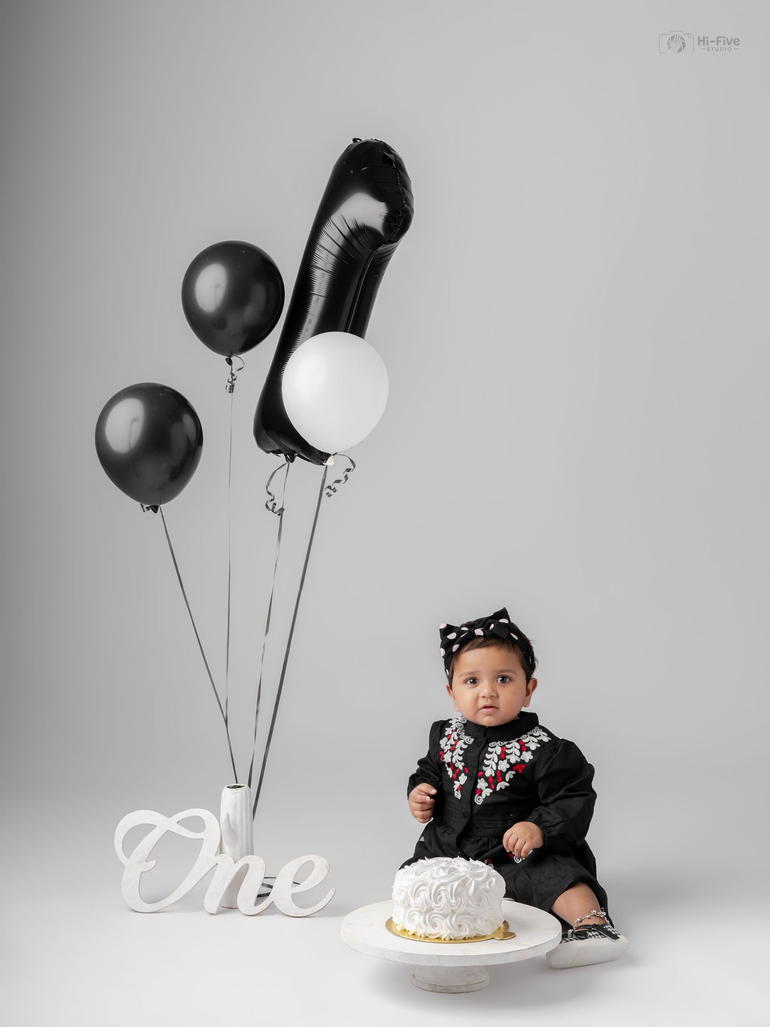 Baby in black outfit sits beside a white smash cake and black balloons for a first birthday photo shoot.