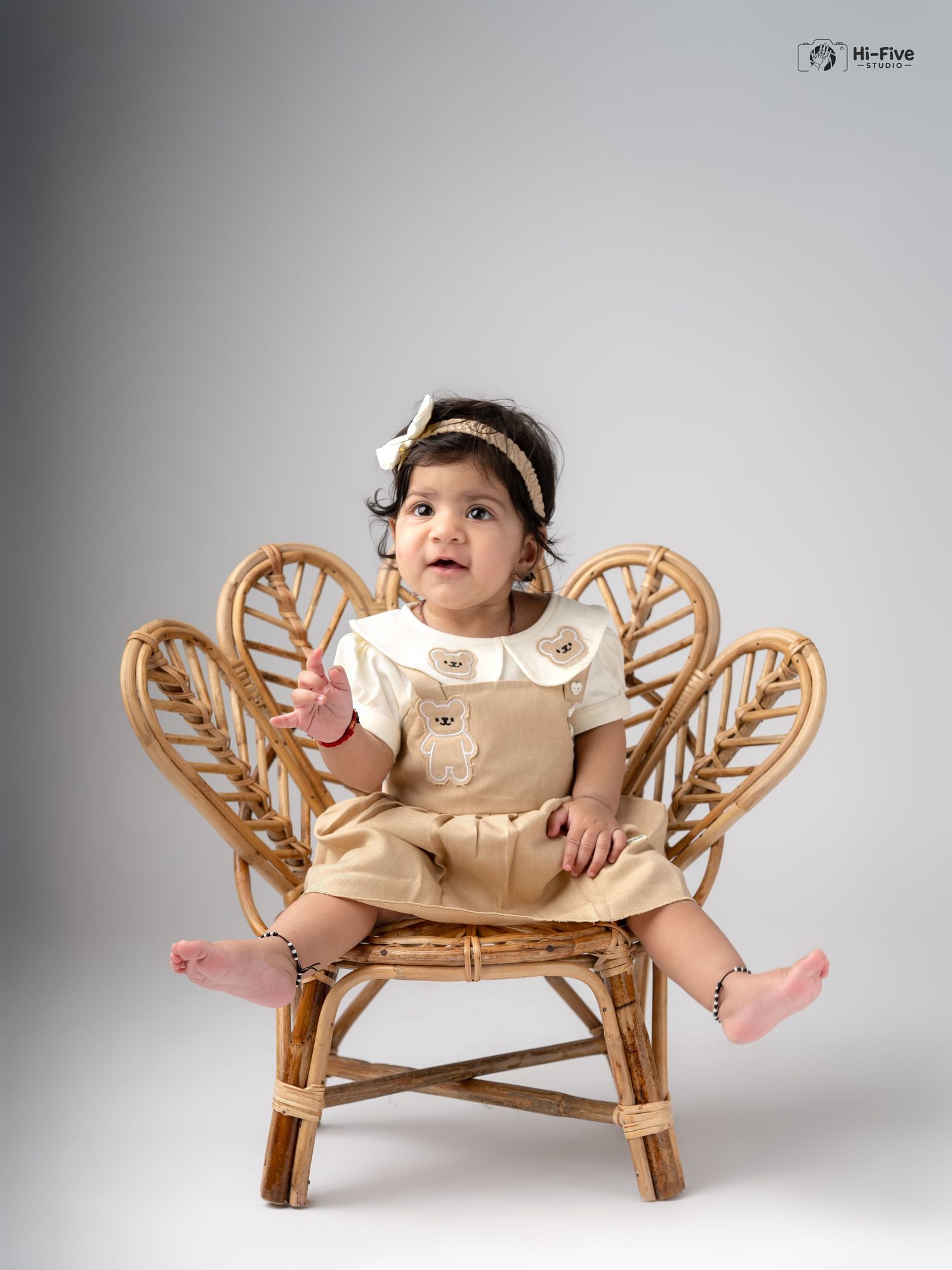 Adorable baby girl in tan dress on wicker peacock chair reaching out with curiosity.