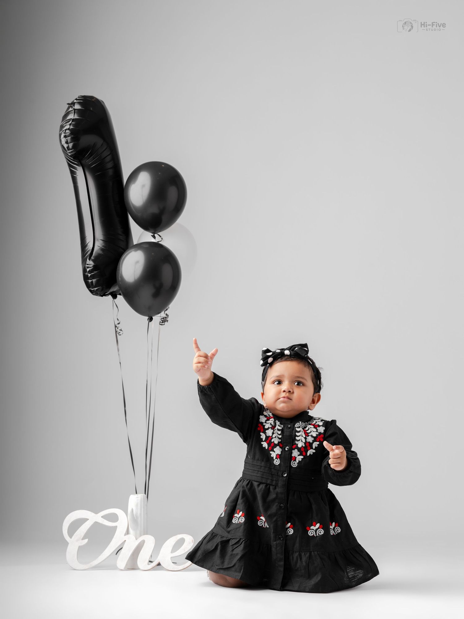Baby girl in black floral dress poses with black number one balloons celebrating her first birthday.