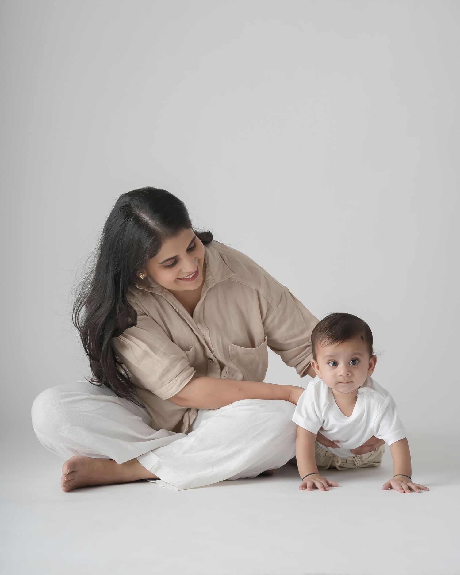 A woman in beige shirt and white pants sits cross-legged on white floor smiling at crawling baby in white onesie.