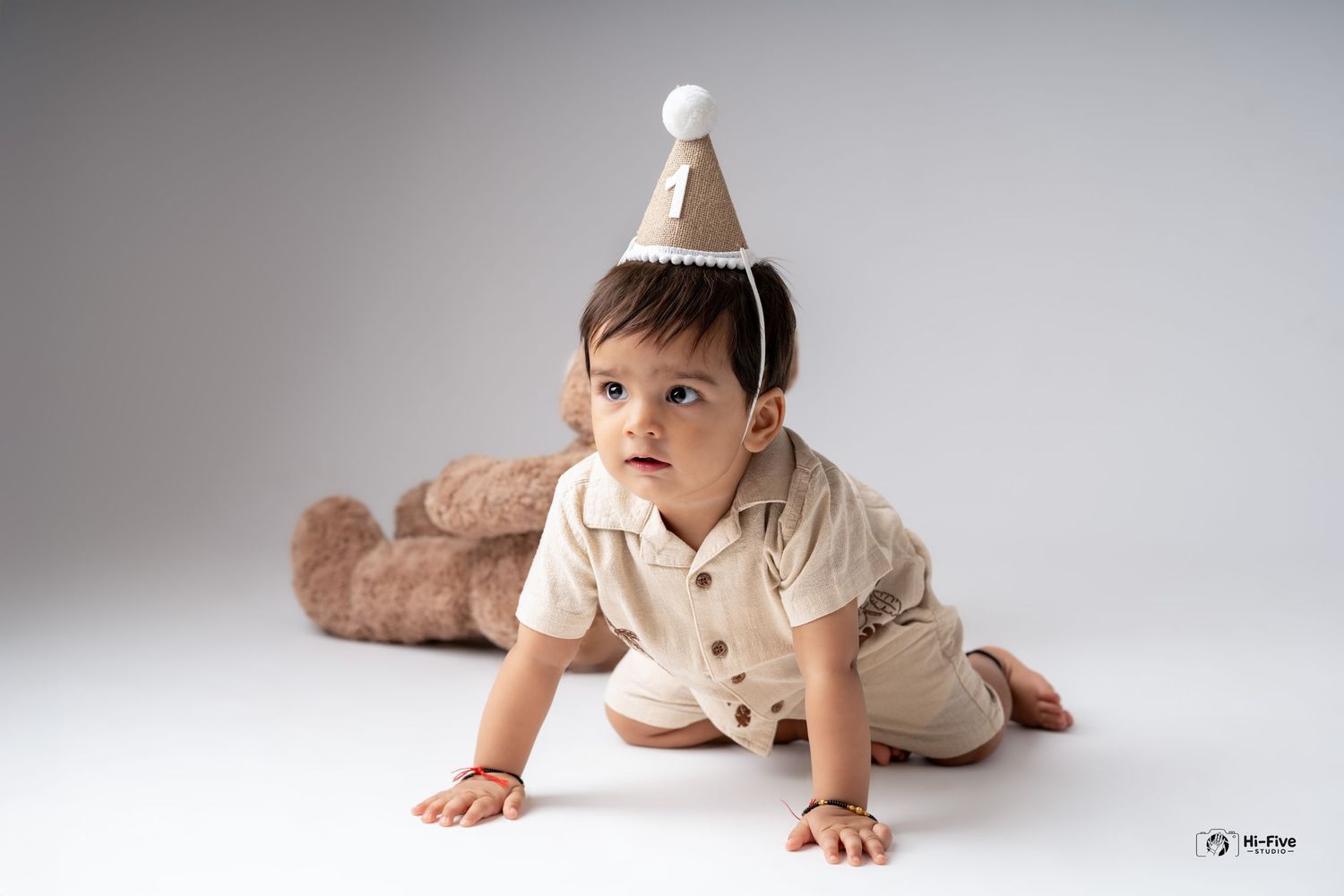 A baby boy wearing a birthday hat crawls on the floor with a teddy bear in the background.