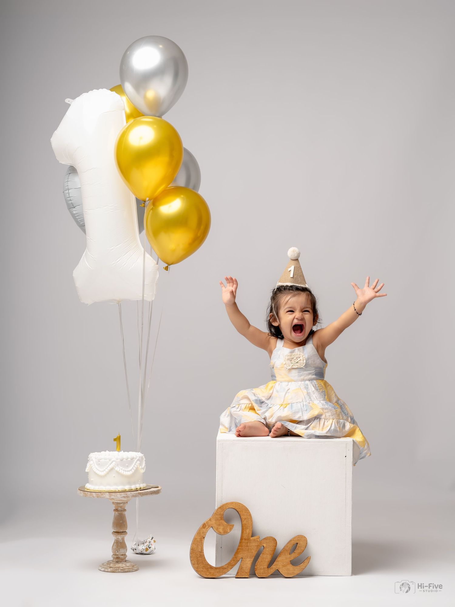 Joyful baby girl celebrates first birthday with gold balloons, white cake, and party hat in a bright studio photo.