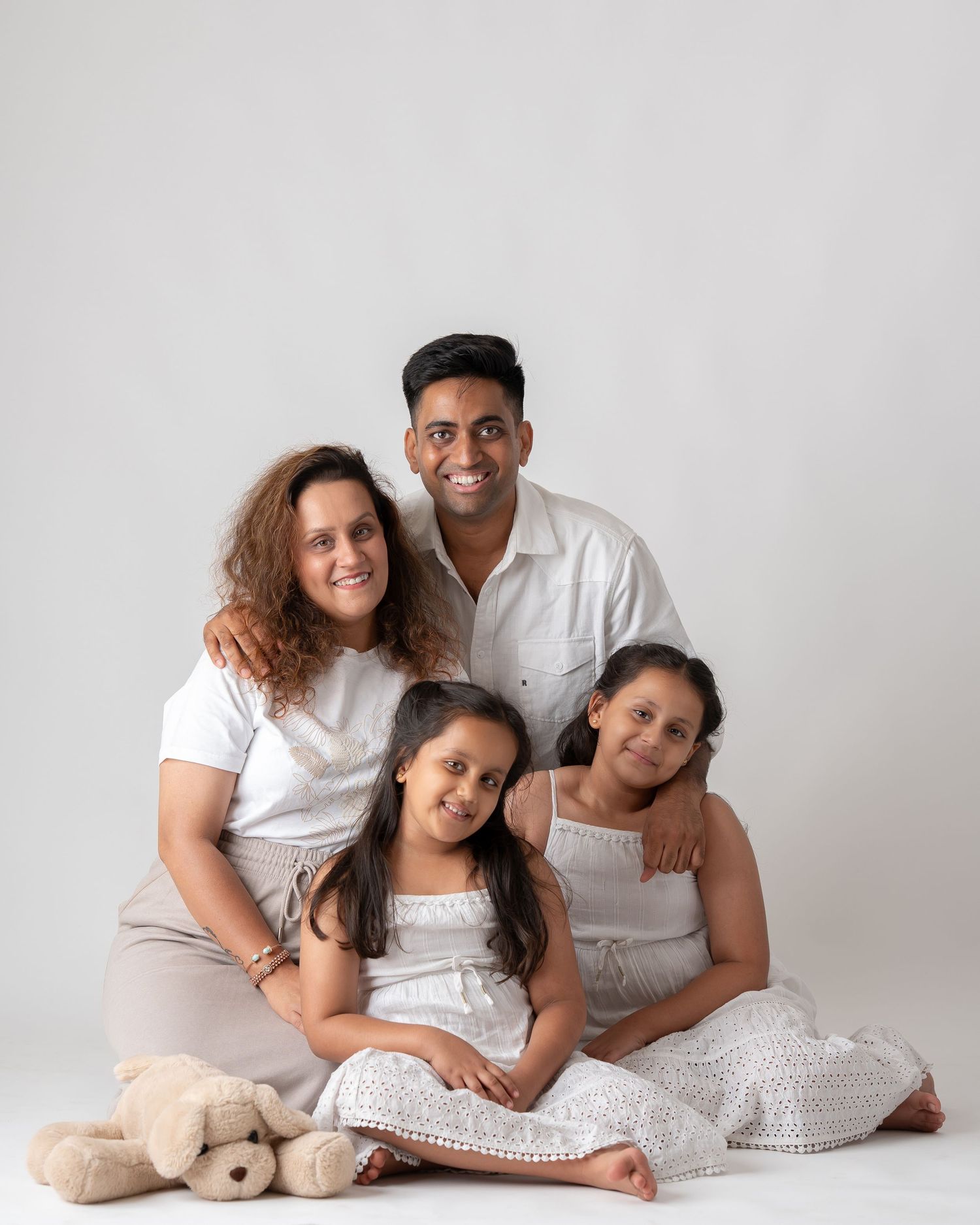 Family of four with two girls in white dresses posing with a teddy bear in a studio portrait.