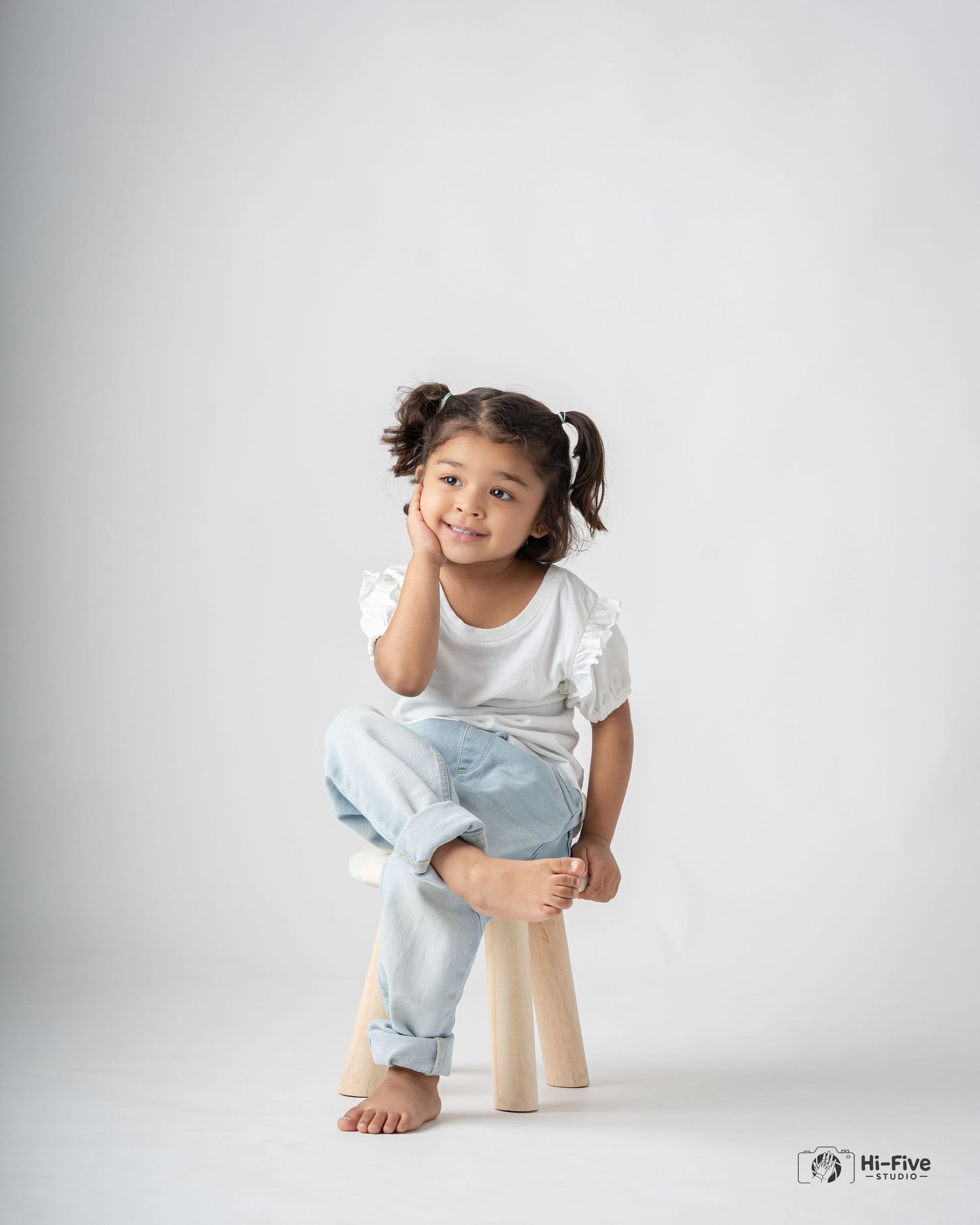 Adorable young girl with pigtails sitting on wooden stool in white top and light jeans.