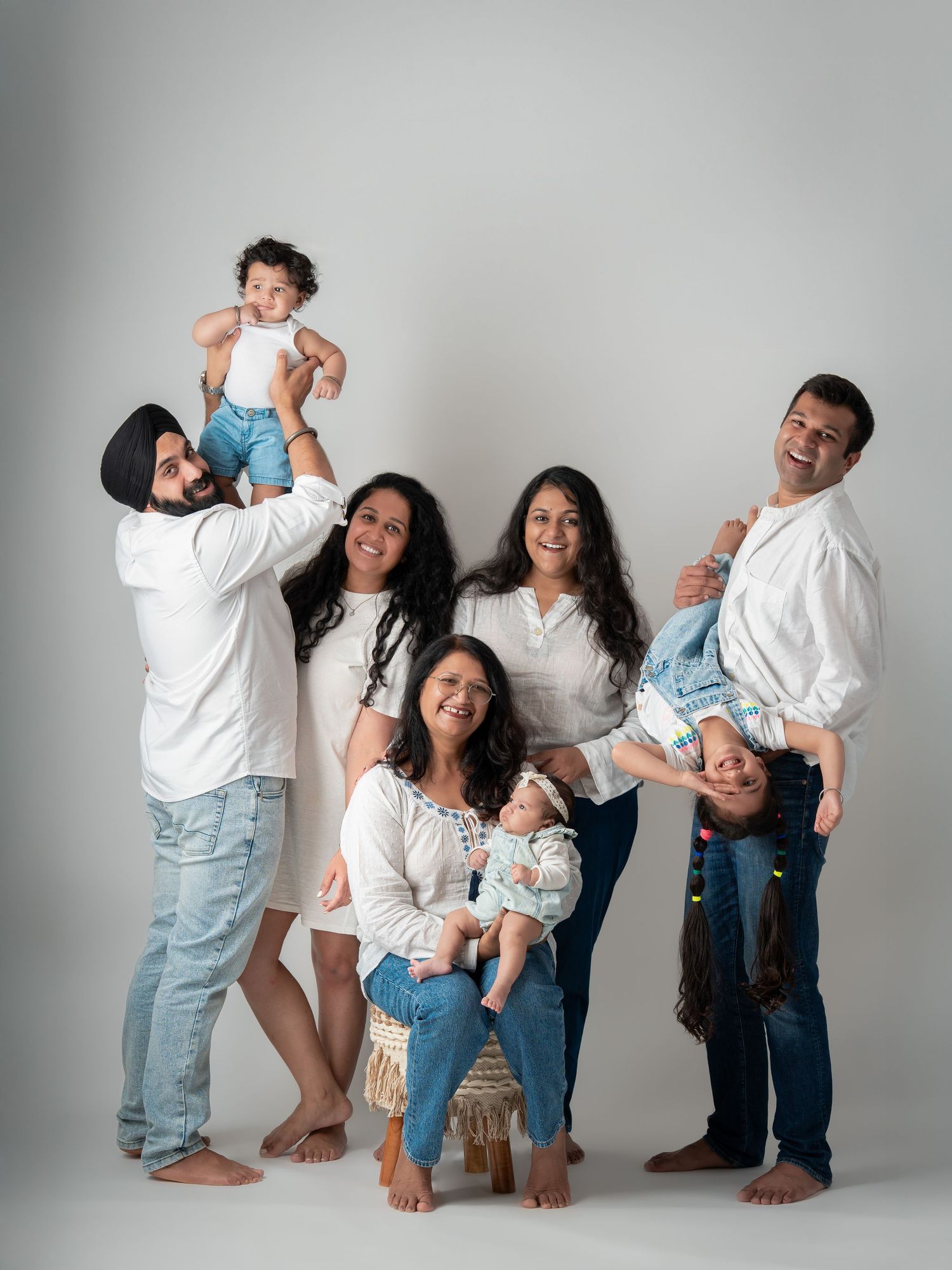Joyful extended South Asian family portrait in white and denim, featuring adults and children in a playful studio setting.