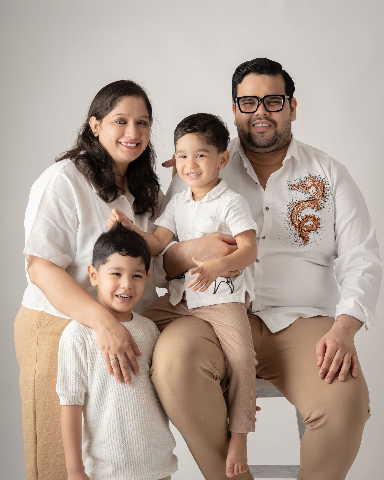 Family of four in matching white and beige outfits posing for a studio portrait with two young boys.