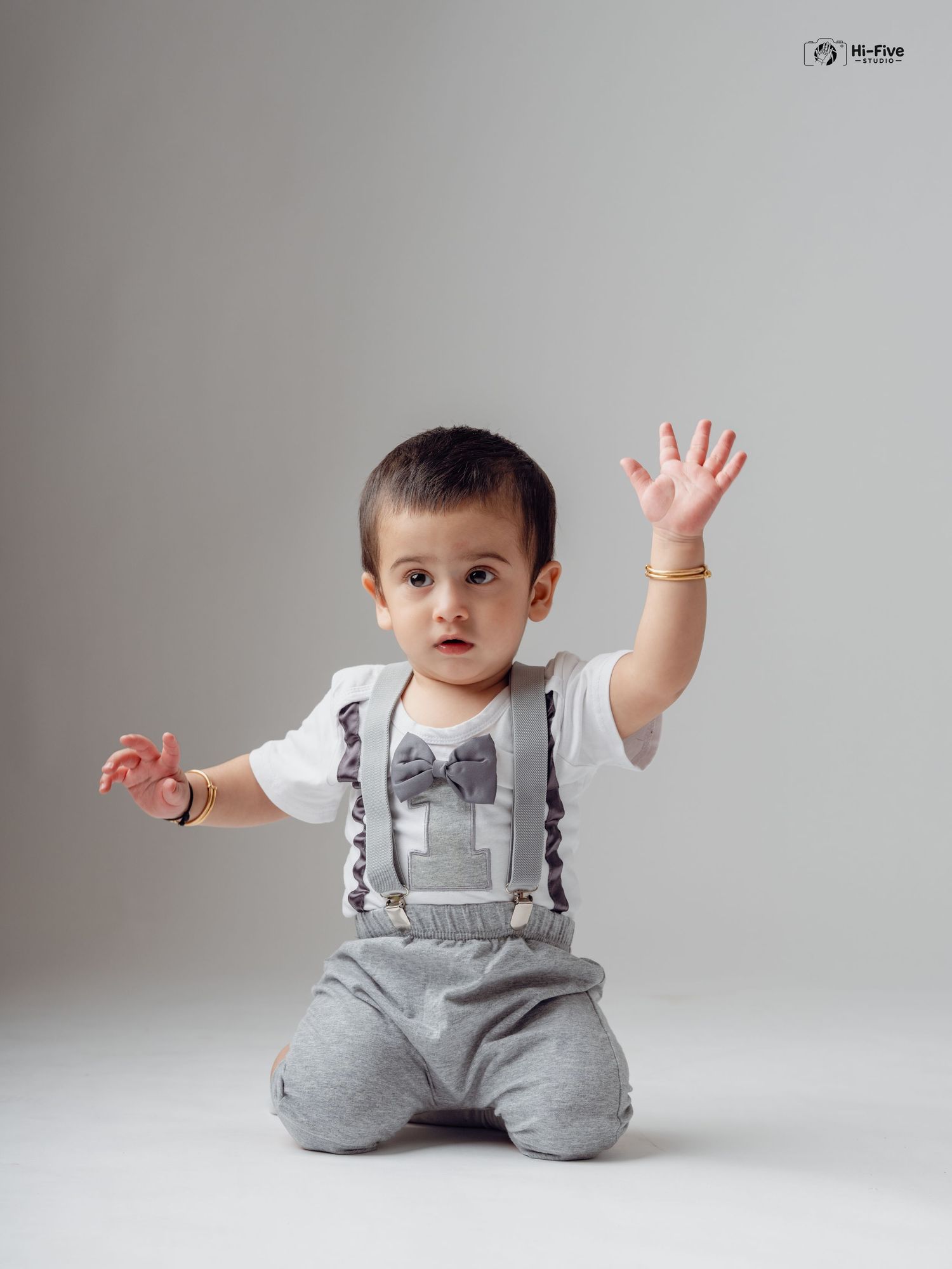 Cute baby boy in gray suspender outfit with bow tie sitting on floor waving hand at camera.