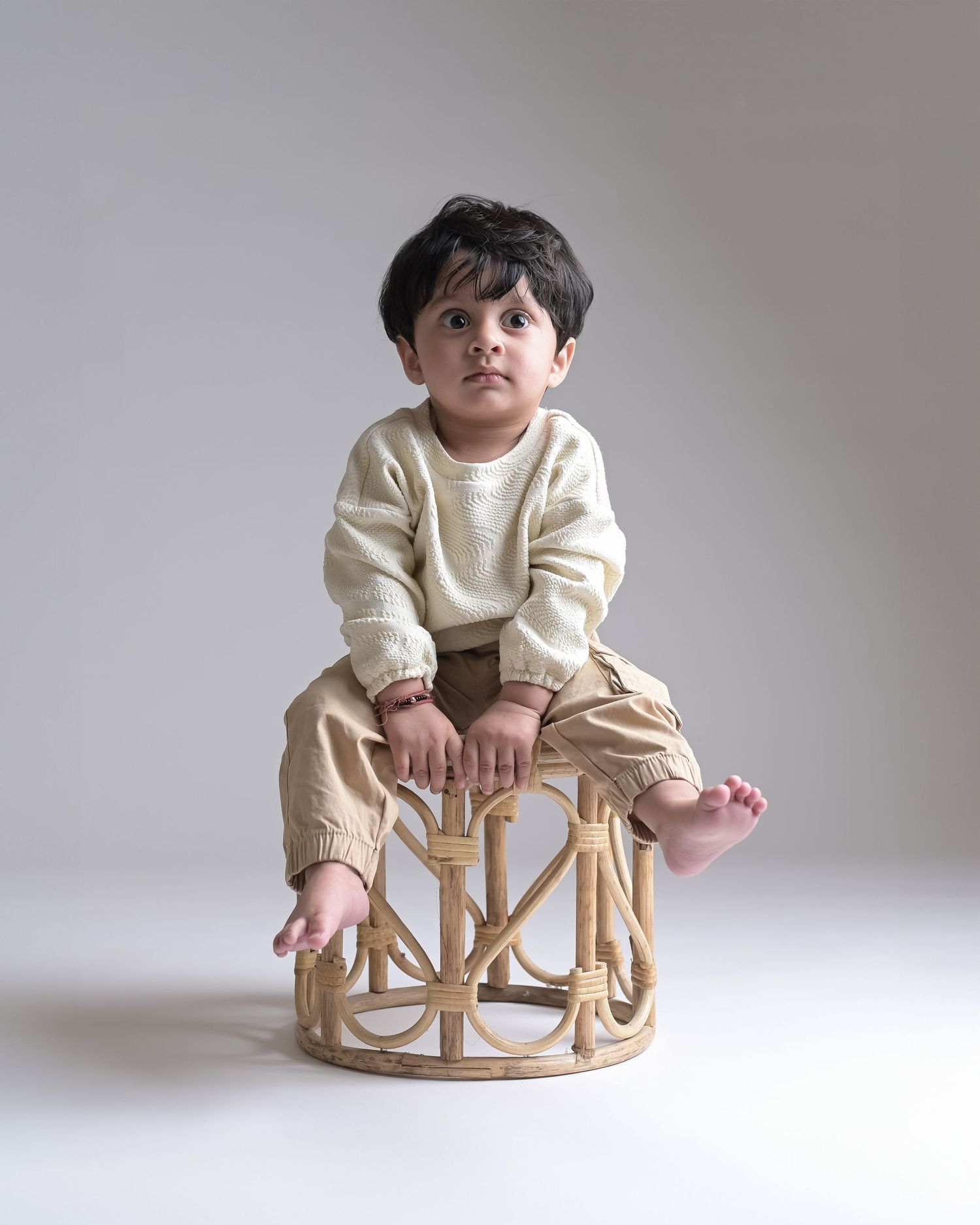 Young toddler boy sitting on rattan stool in cream sweater and beige cargo pants.