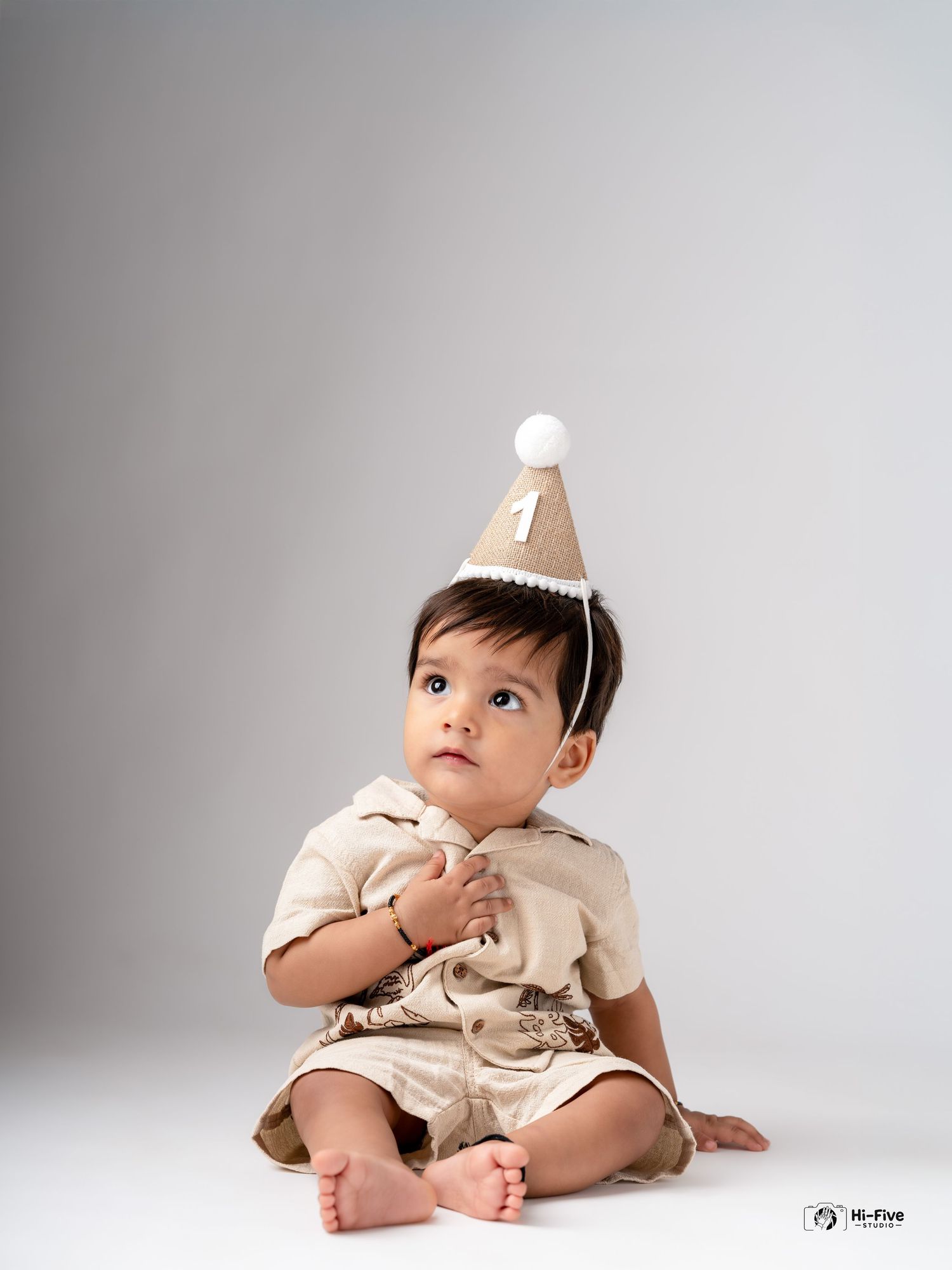 A cute baby boy wearing a number one birthday hat sits on the floor in a studio photo.