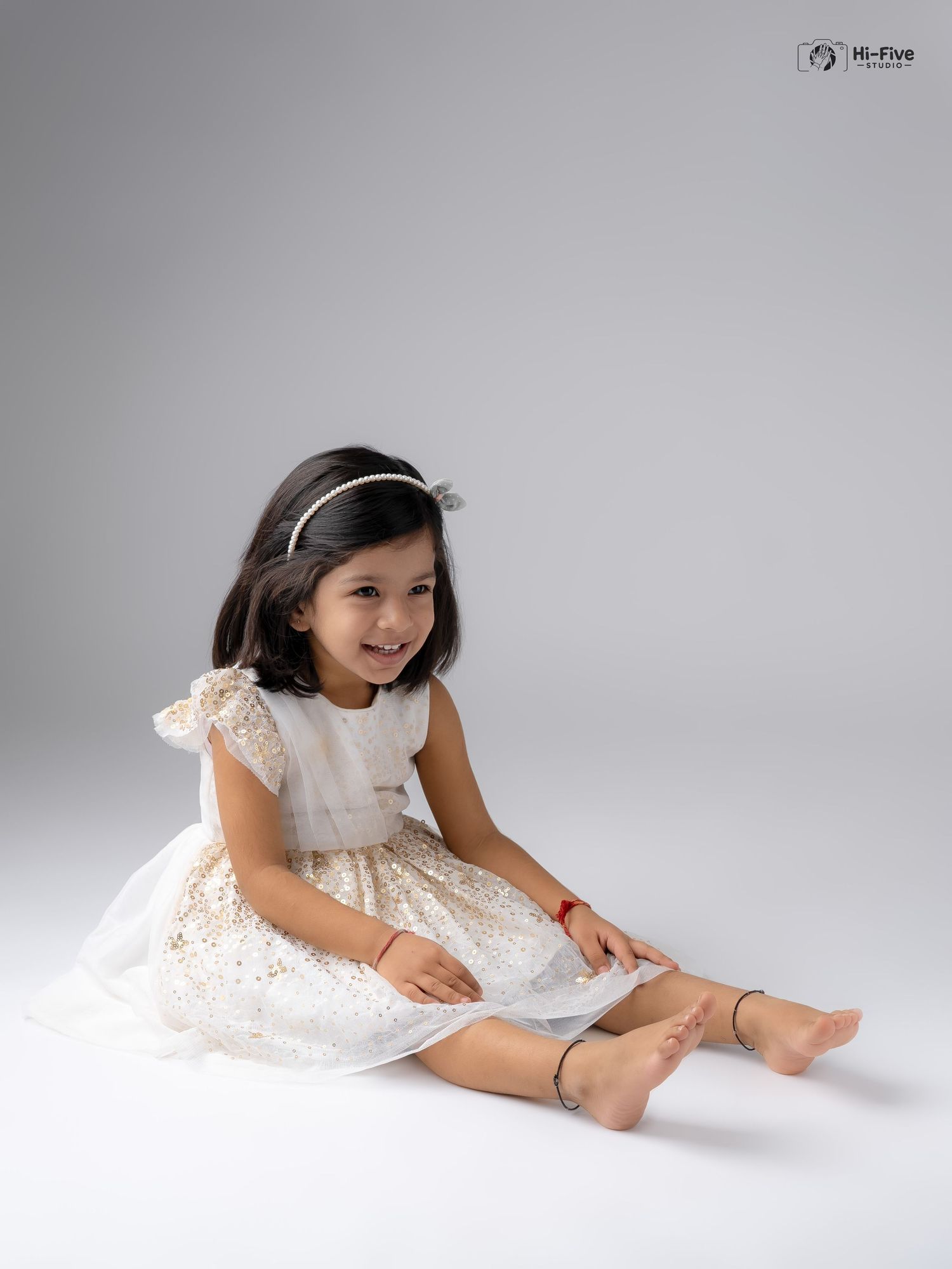 Happy young girl in white sparkle dress smiles while sitting on floor during professional photoshoot.