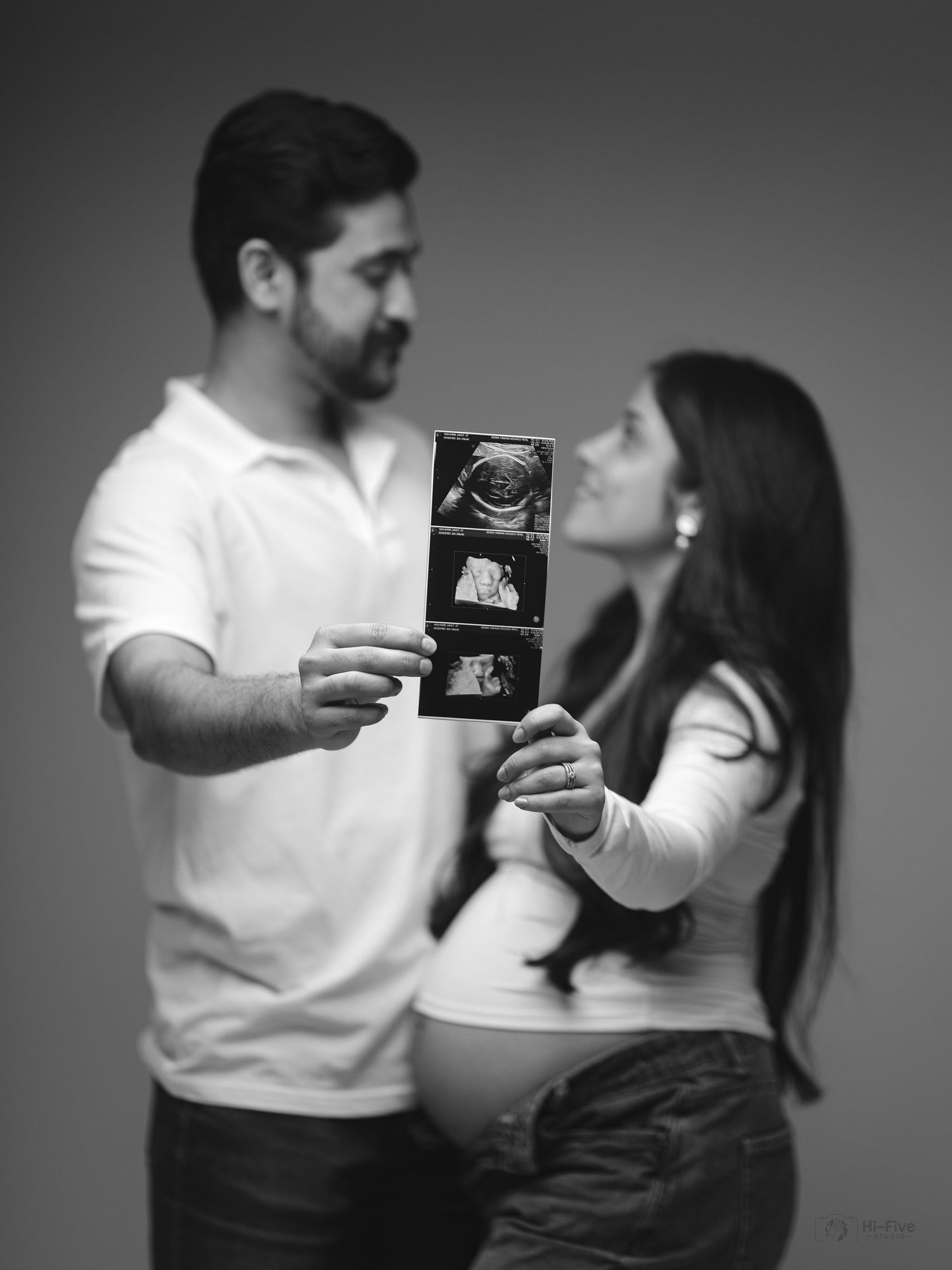 Couple holding ultrasound photos in black and white maternity portrait with pregnant belly visible.
