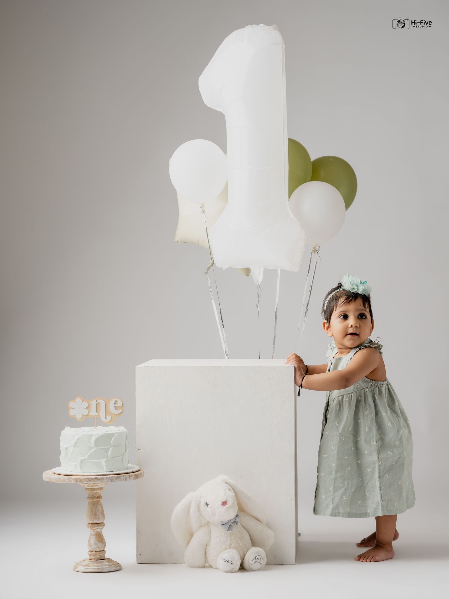 Baby girl in sage green dress poses with number one balloon, bunny toy, and birthday cake in studio.
