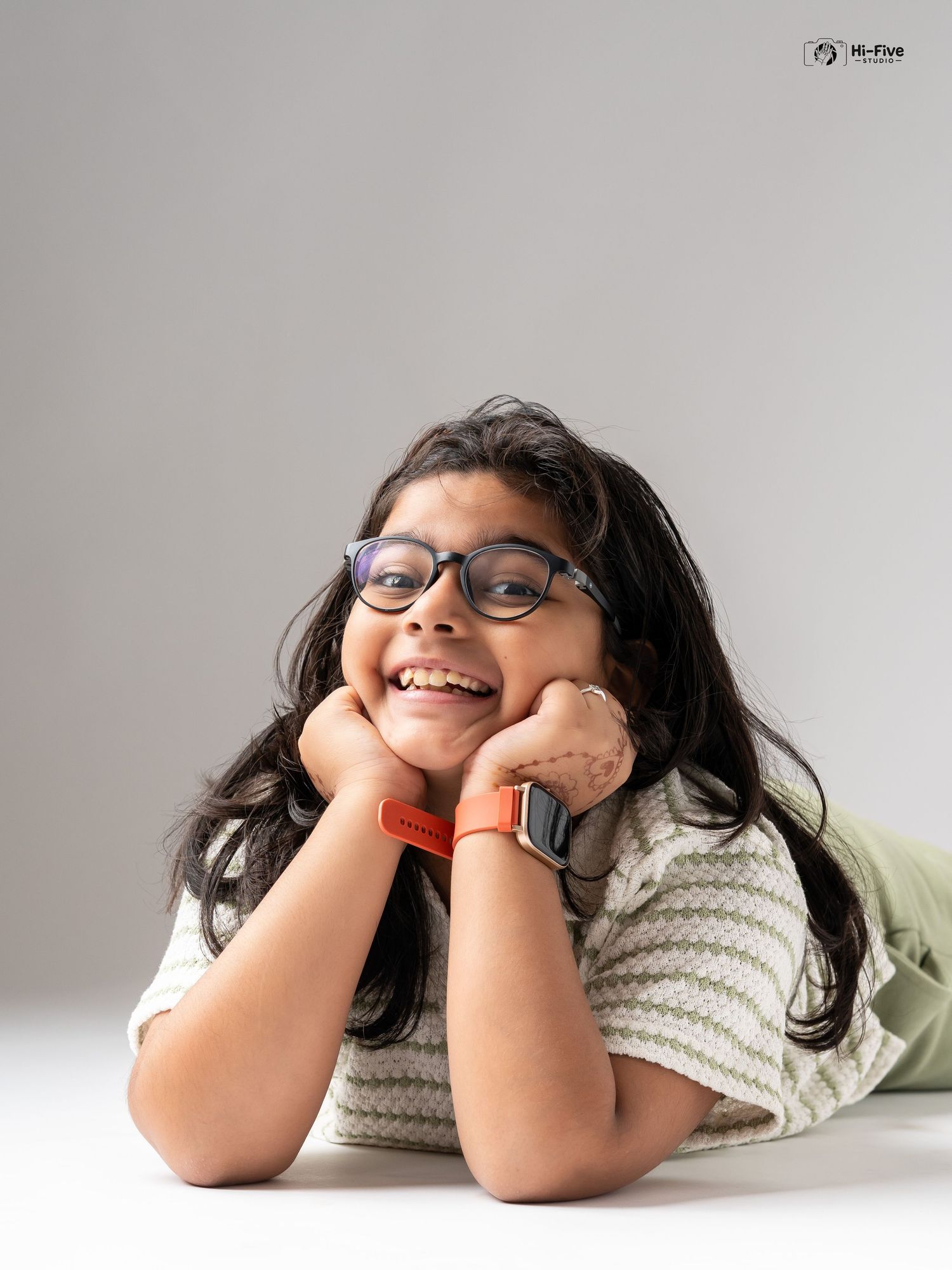 A joyful young girl with glasses lies down smiling broadly, resting her chin on her hands, wearing a striped top.