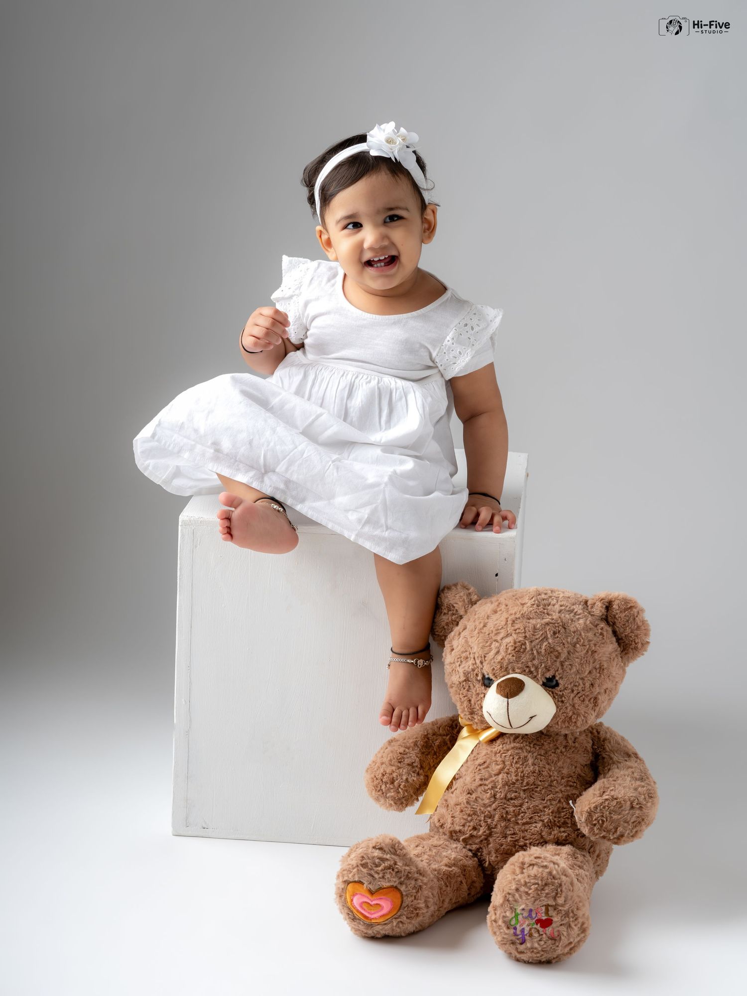 Happy baby girl in white dress sitting on white cube next to large brown teddy bear.