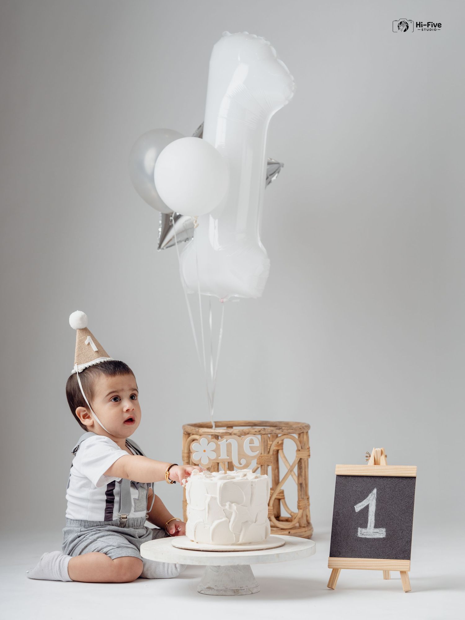 Baby boy in suspenders touching first birthday cake with number one balloons and chalkboard sign.