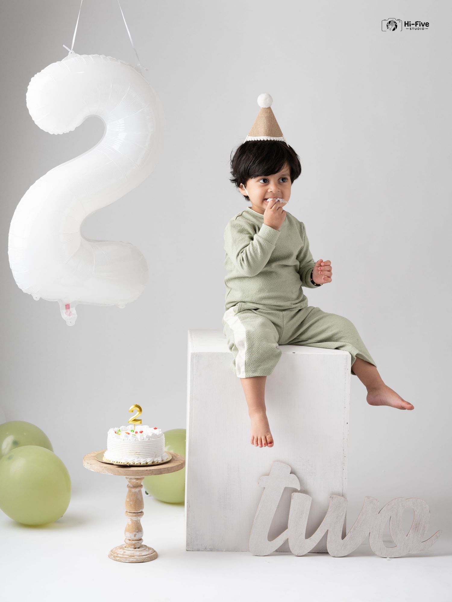 A toddler in a green outfit and party hat sits on a white cube beside a birthday cake celebrating turning two.