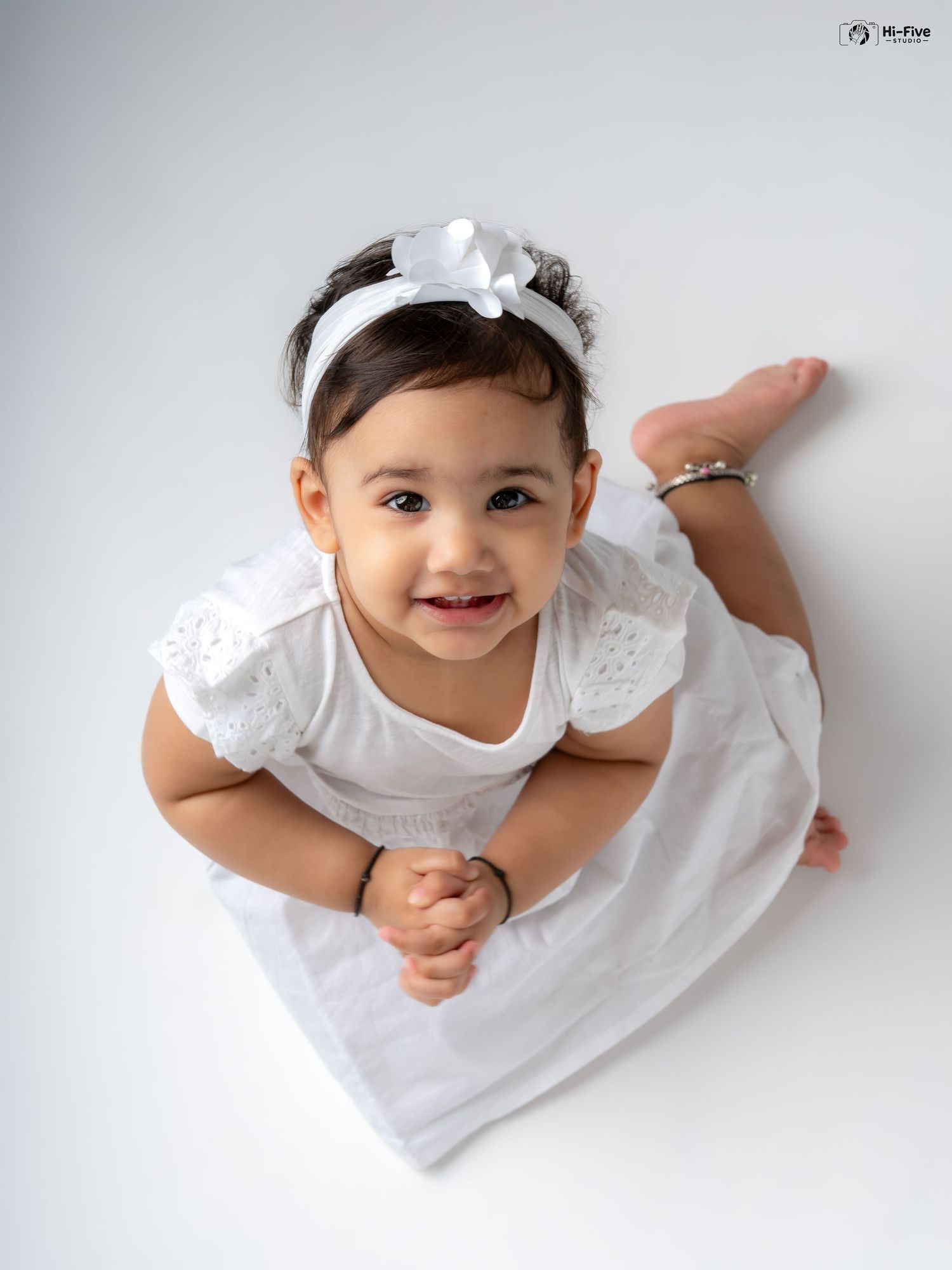 Cute baby girl in white dress lying on tummy smiling, wearing floral bow headband.