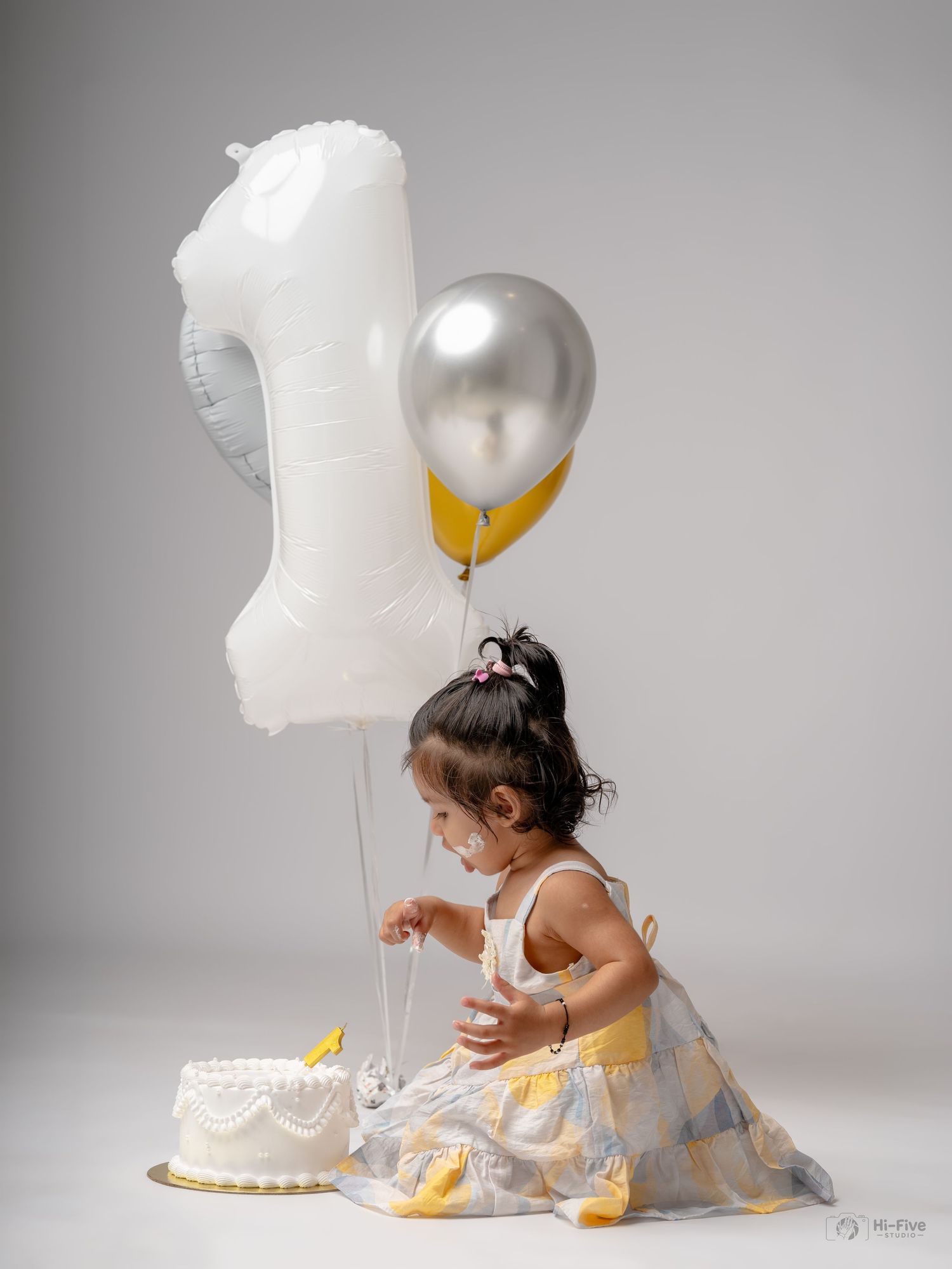 A toddler girl in a yellow dress sits beside a first birthday cake with number 1 balloon and silver balloons behind her.