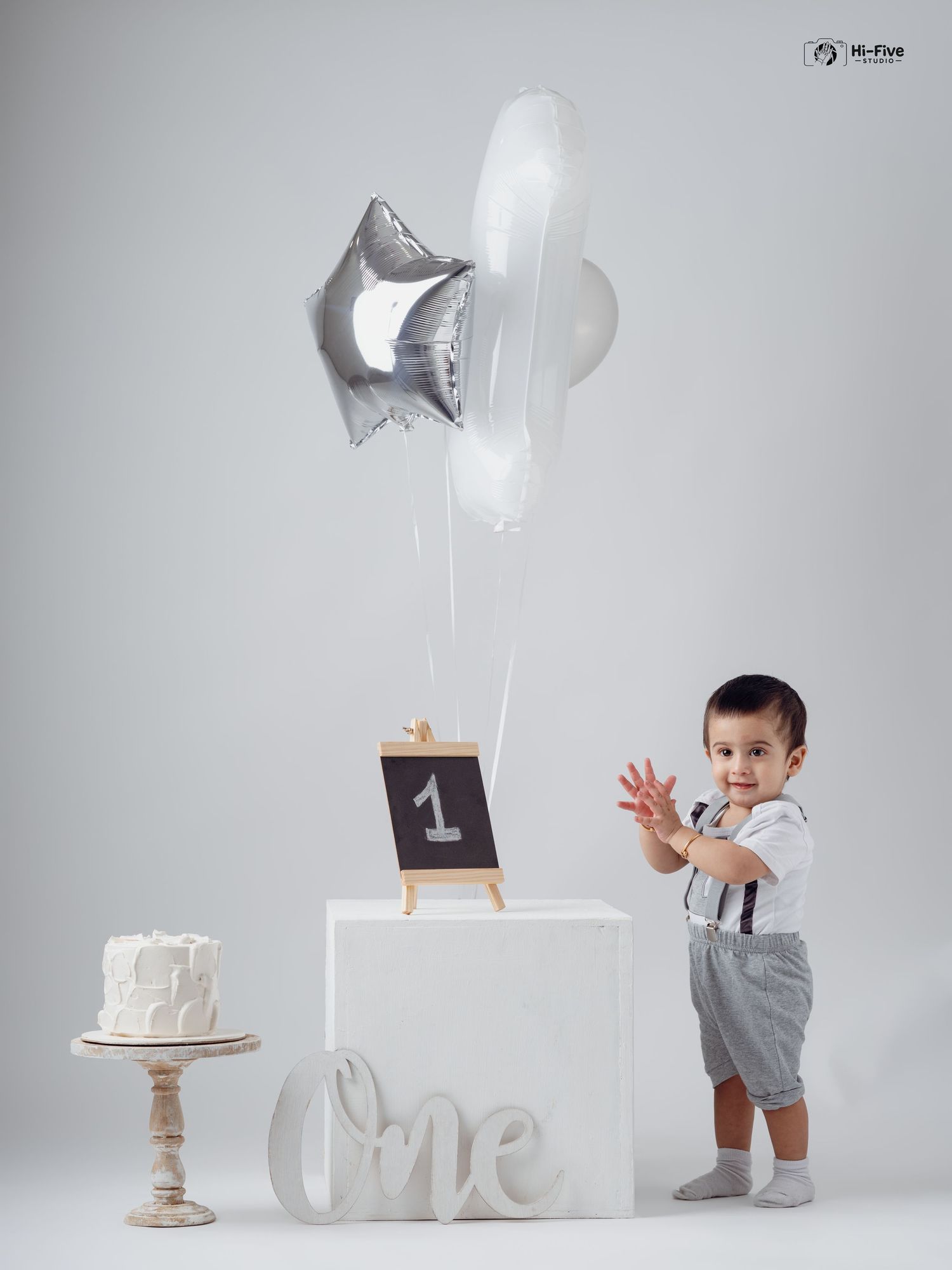 A toddler boy celebrates his first birthday with a cake, balloons, and one sign in a studio.