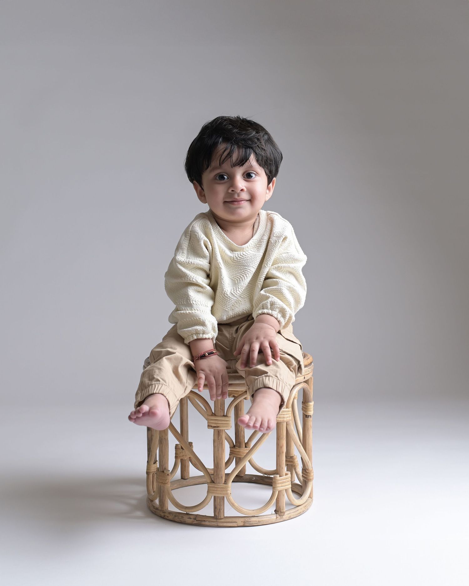 Cute toddler boy smiling on woven rattan stool in neutral cream and tan outfit.